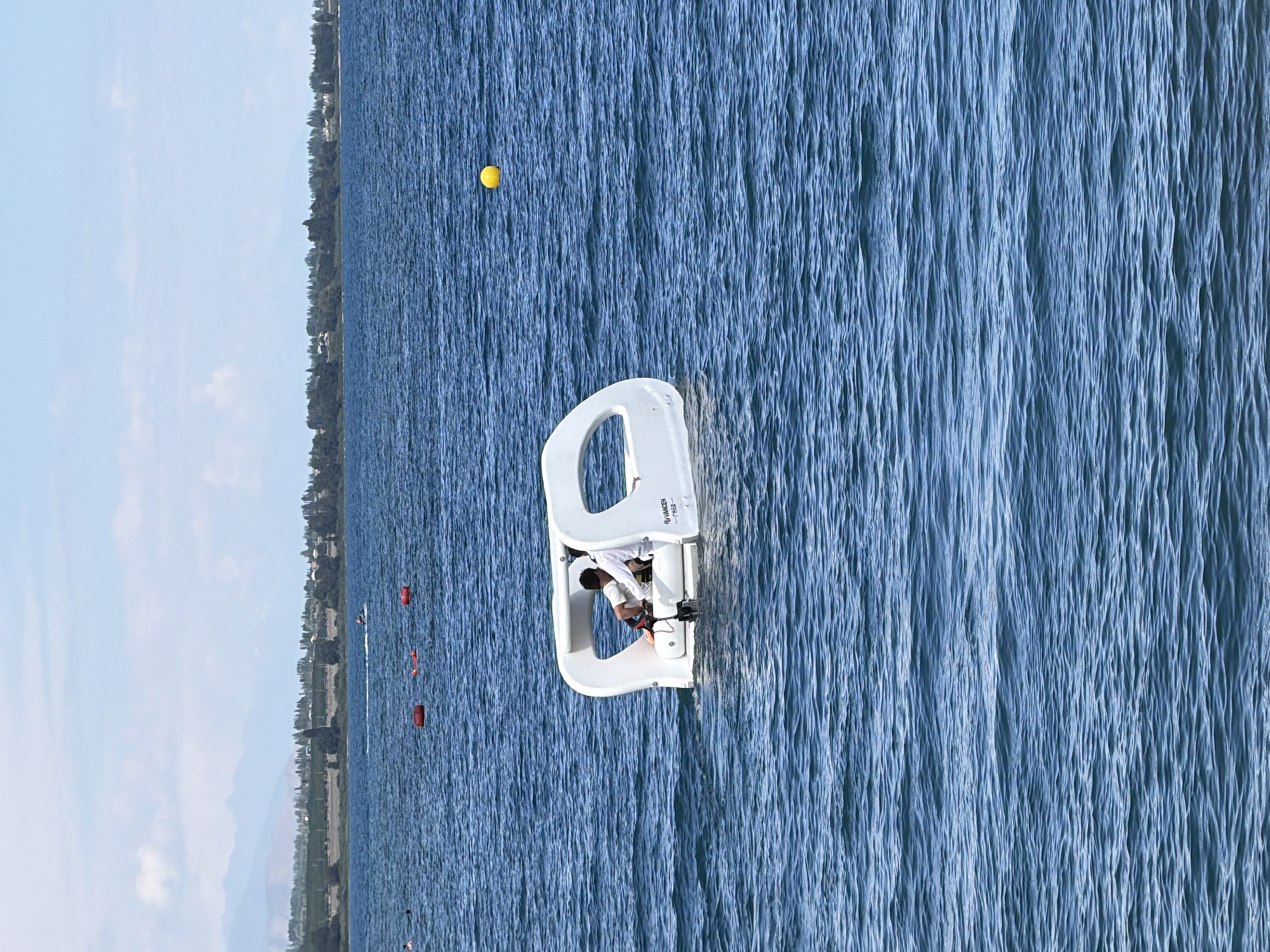 This media file displays a single, static shot of a person on a small, white boat. The boat appears to be a pedal boat, with an open design and a seating area for at least one passenger. The person in the boat is wearing a white shirt and dark shorts. They are seated in the boat, with their hands on what seem to be pedals. The boat is on a large body of water, likely a lake or the sea, with the water's surface rippled by wind. In the distance, a shoreline is visible, lined with trees and some buildings. The sky is a light blue with some scattered clouds. A yellow buoy is visible in the water to the left of the boat. A few red buoys are also visible in the distance. The image is oriented vertically. There is some text on the boat, but it is too small to read. The scene suggests a recreational activity, possibly on a sunny day.