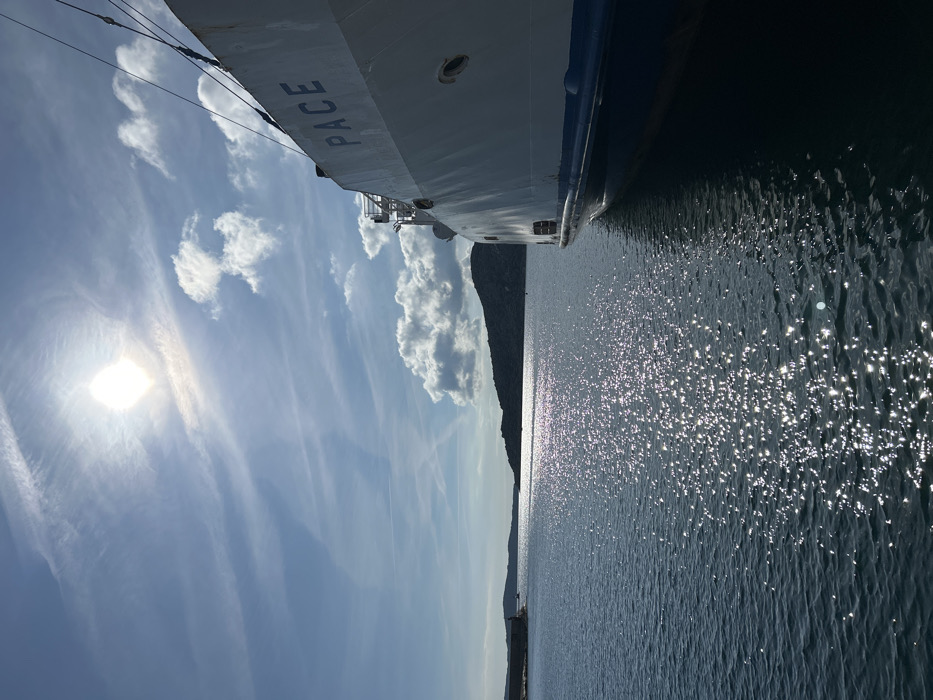 The scene depicts a bright, sunny daytime view of a harbor in La Maddalena, Italy. A large white vessel, identified by the name "PACE" in blue lettering on its hull, occupies the right side of the frame, appearing stationary or docked. The ship's hull features a blue stripe along the waterline and several circular openings. The sun, positioned in the upper left, is intensely bright and casts a shimmering path of specular reflection across the calm, dark blue water. The sky is predominantly clear blue with scattered white cumulus clouds and faint wispy cirrus clouds, including visible aircraft contrails. In the midground, dark landmasses, consistent with islands or distant coastlines, are visible along the horizon. To the far left, a concrete or stone breakwater structure marks the entrance to a narrower body of water. No individuals are visible, and no specific actions or interactions are in progress.