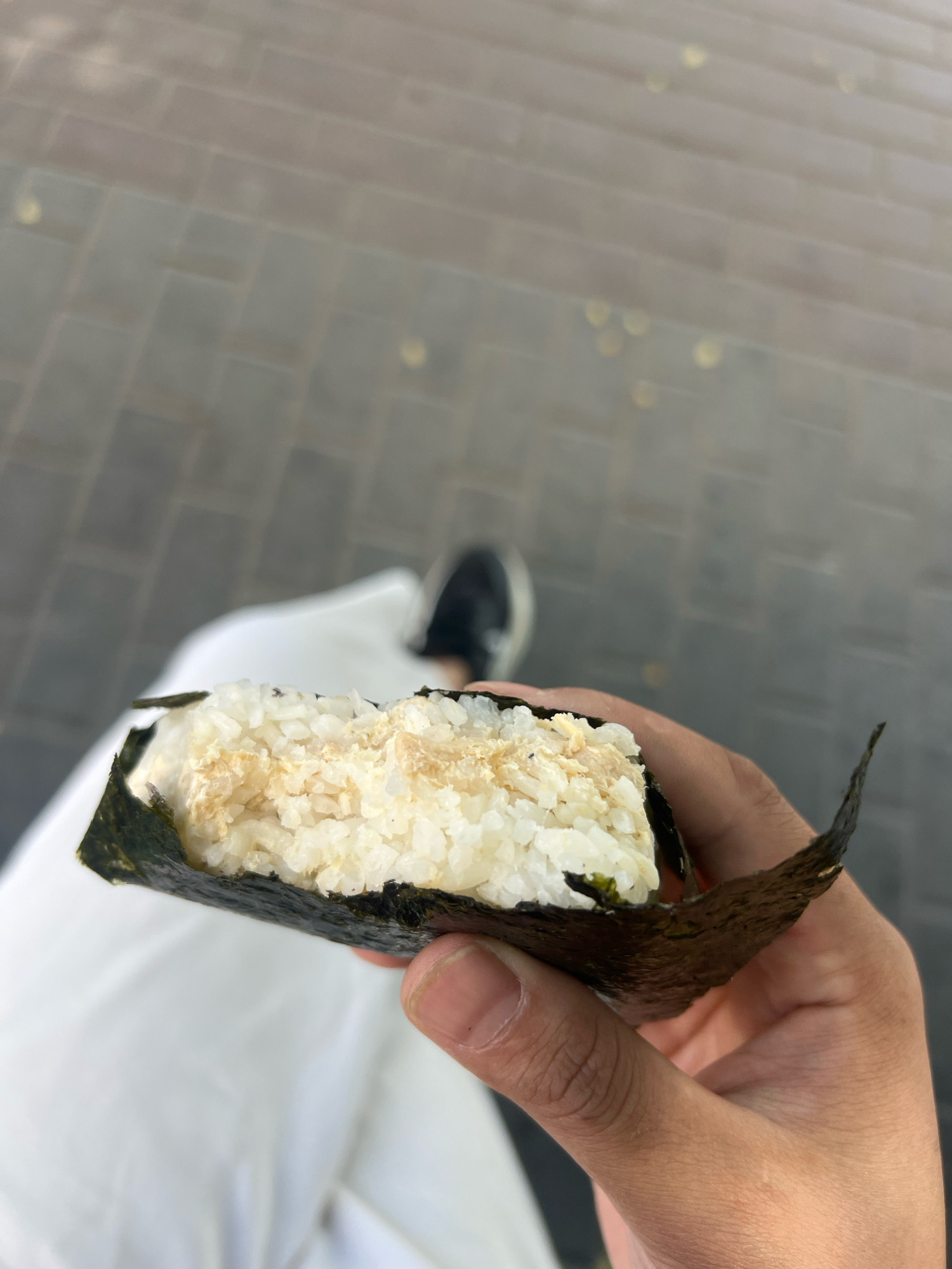 A person's hand holds a triangular onigiri, a Japanese rice ball wrapped in seaweed, outdoors. The onigiri has white rice and a filling that appears to be shredded chicken or tuna mixed with mayonnaise. In the background, a person is wearing white pants and dark sneakers, walking on a paved walkway. The walkway is made of rectangular stone pavers, some of which are illuminated by overhead lights, suggesting it might be evening or a dimly lit area. The overall environment appears to be an urban setting in Osh City, Kyrgyzstan.