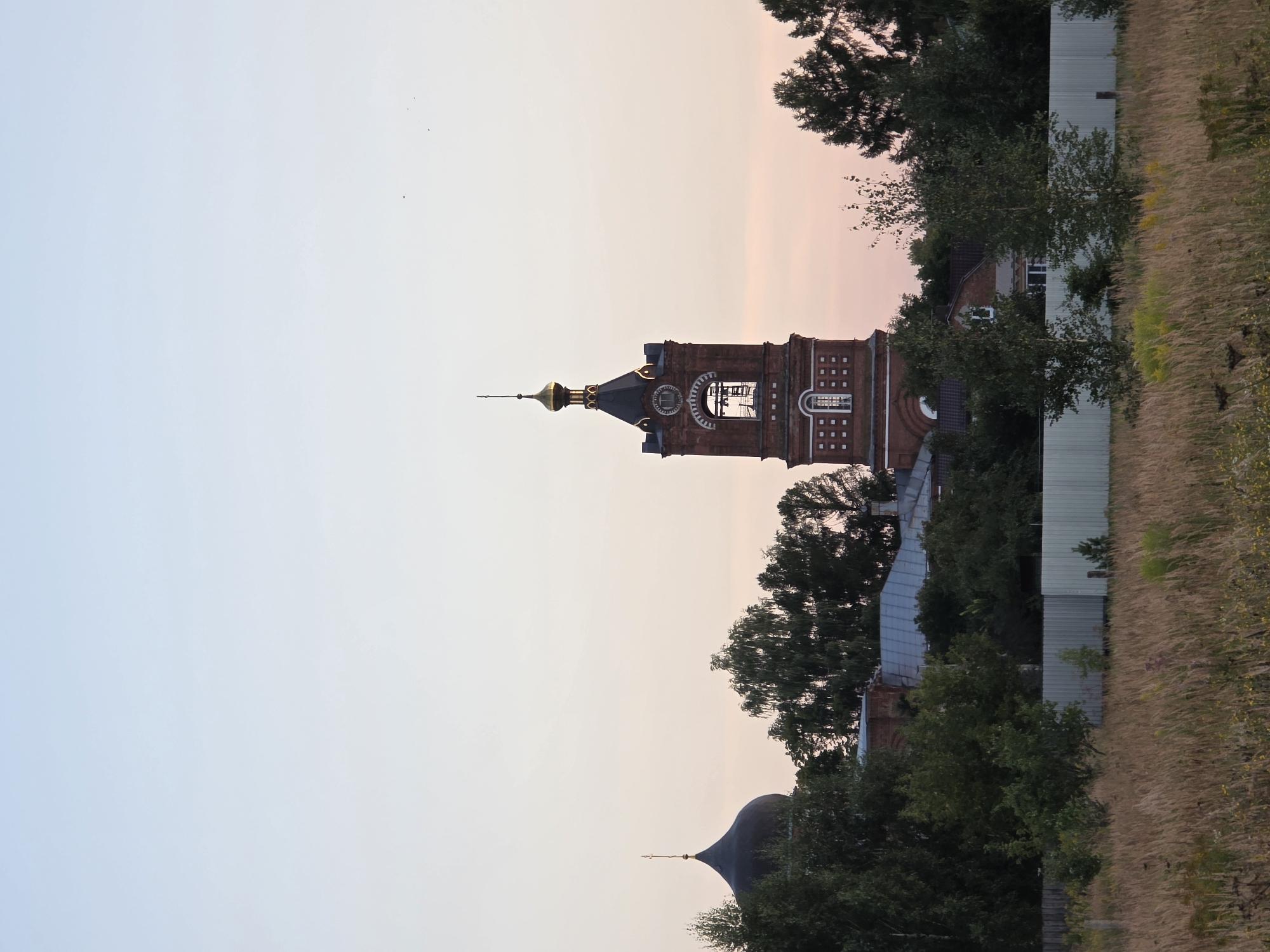 A distant view of a red brick church with a golden dome and a tall bell tower is captured at dusk. The church is situated amongst lush green trees, and a small, dark dome with a cross is visible to the left of the bell tower. In the foreground, a field of dry grass stretches towards a light-colored fence. The sky displays a gradient of pale blue to soft pink, indicating the transition from day to night. The scene appears to be located in the city of Rechitsy, Russia. There are no people or animals visible in the image.