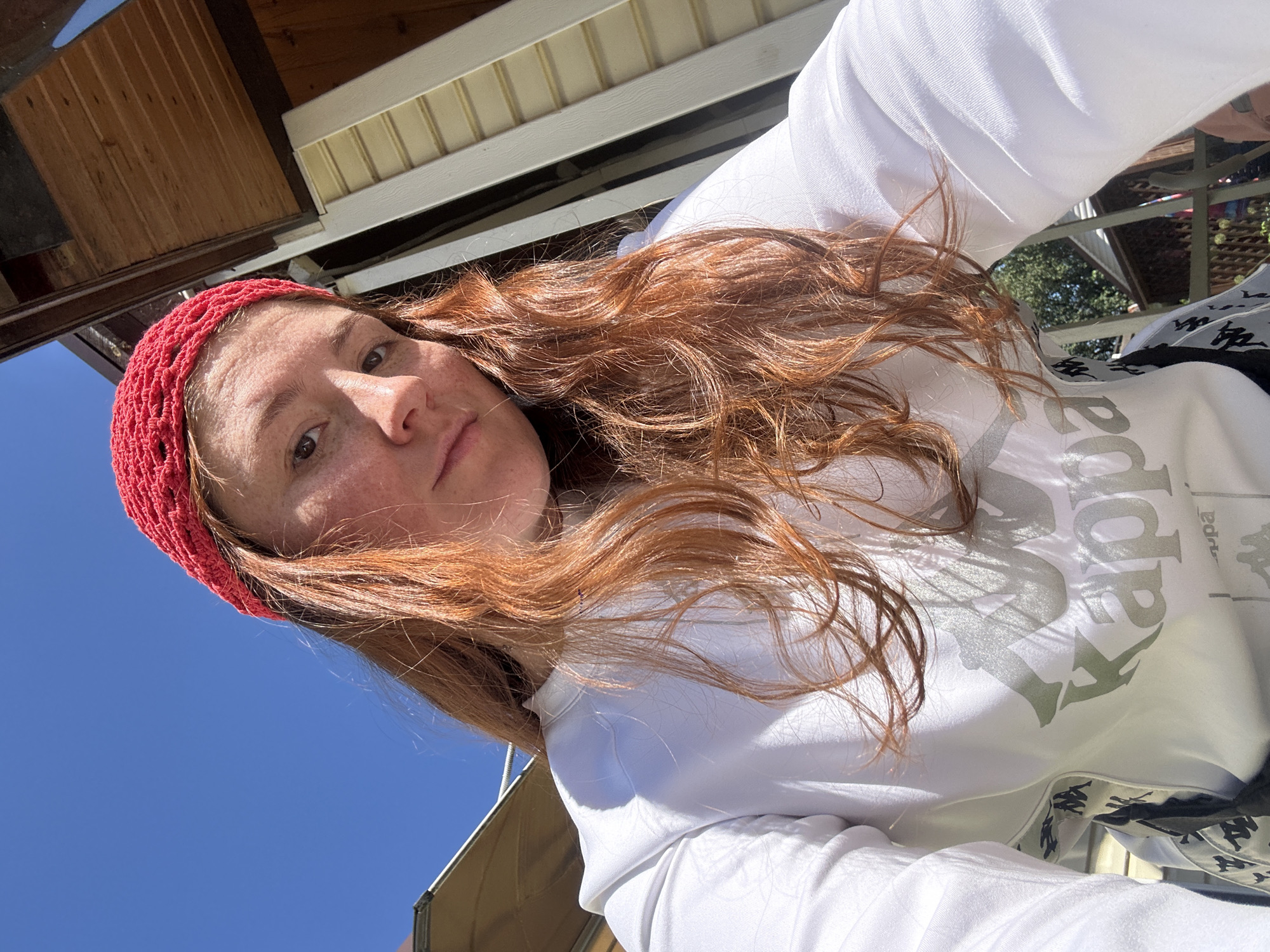 A young person with long, wavy, red hair, wearing a red crocheted beanie and a white sweatshirt with a Kappa logo, is photographed from a low angle, looking up at the camera. They have freckles on their face. The background consists of a bright blue sky, a wooden structure that appears to be an overhang or part of a building, and some greenery. The lighting suggests it is daytime and sunny. The image does not contain any information or visual cues that would allow for its location to be identified as Nizhny Novgorod, Russia, or any specific city.