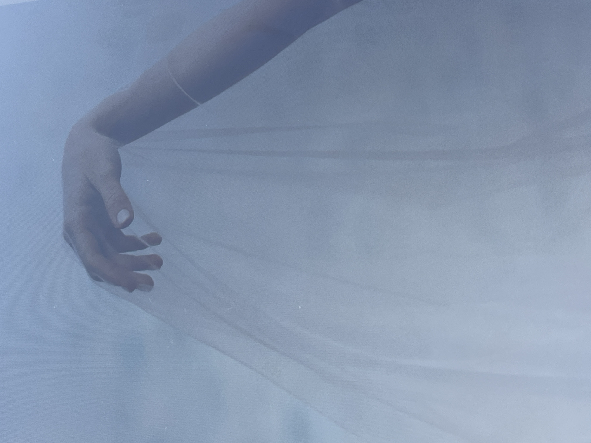 A photograph shows a person's arm and hand partially obscured by a translucent white fabric. The hand is positioned on the left side of the frame, with the fingers slightly spread. The arm extends upwards from the hand, disappearing at the top of the frame. The background appears to be a muted blue-gray. The location is unknown.