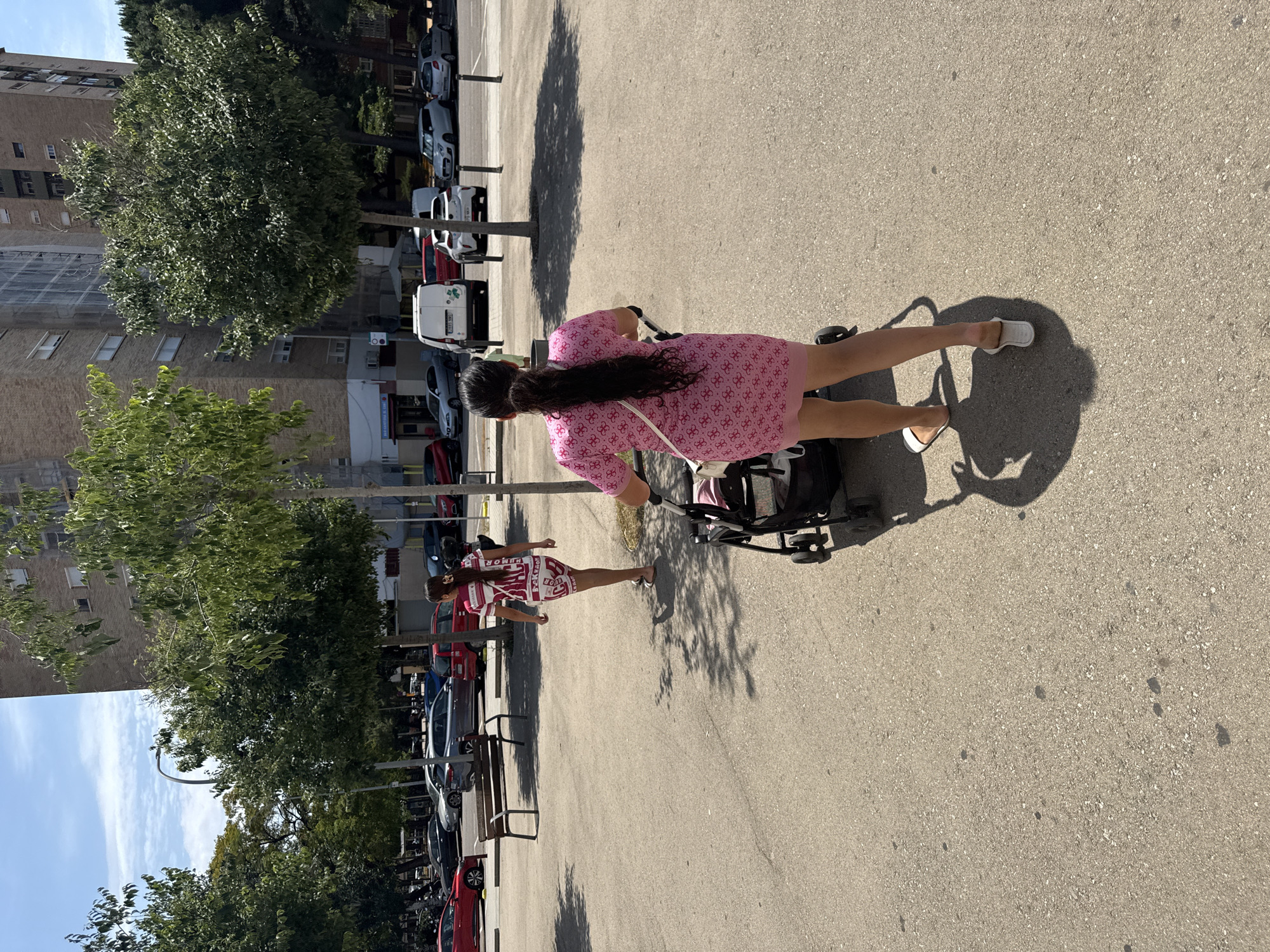 In L'Hospitalet de Llobregat, Spain, two women are seen walking in an outdoor urban environment on a sunny day. The woman in the foreground, facing away from the camera, is pushing a stroller. She is wearing a pink dress with a floral pattern and white flip-flops. The stroller is dark-colored with black wheels. Behind her, another woman, also facing away, is walking further ahead. She is wearing a white and red patterned dress and sandals. The background features a tall, multi-story building with many windows, several trees with green foliage, and a parking lot with various cars. The ground is paved with asphalt.