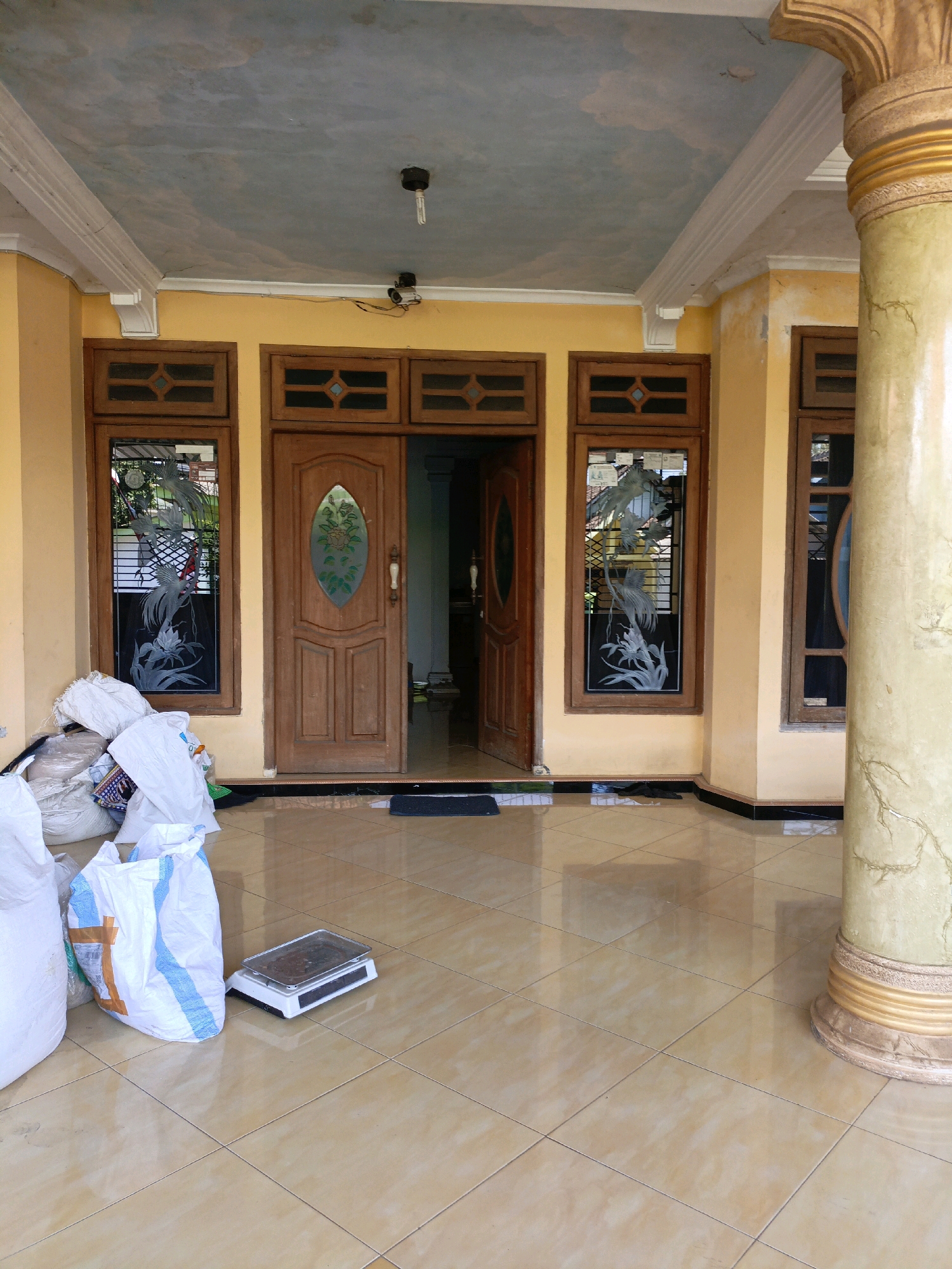 The image depicts the exterior of a building in Betek, Indonesia, focusing on a covered porch area. The porch has a tiled floor and is supported by decorative columns, one of which is prominently featured on the right side of the image. The walls are painted a light yellow. A large wooden double door is the central feature of the entrance, flanked by windows with decorative glass featuring bird and floral motifs. To the left of the entrance, several large white sacks and other bundled items are piled on the floor, suggesting storage or delivery. A digital scale sits on the floor in front of these sacks. The ceiling of the porch is painted with a light blue and grey cloud pattern and has some visible cracks. A light fixture and a security camera are also mounted on the ceiling. The scene is brightly lit, likely by natural daylight. No people are visible in the image.