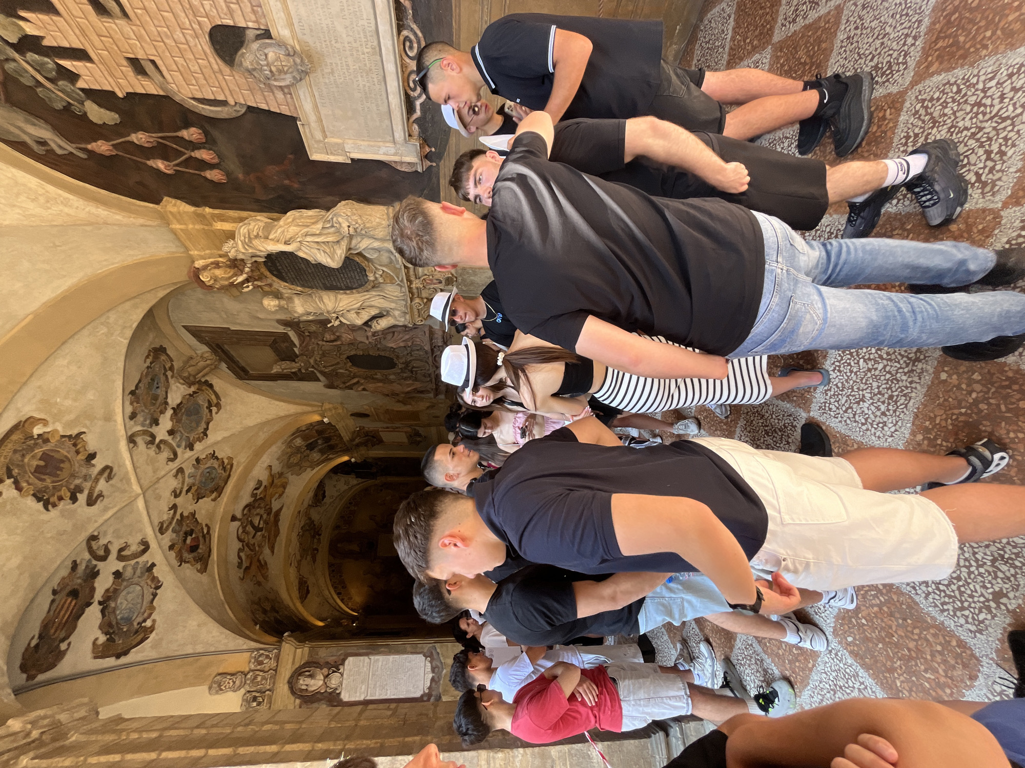 A medium indoor shot shows a row of people looking at a complex, ornamented painted and sculpted ceiling and walls. The location appears to be an arcade in Bologna, Italy. The people are predominantly young, casually dressed, and looking upwards, possibly engaged in a tour or observation of the architecture and artwork. The ceiling features ornate paintings, shields, and decorative elements, while the walls show sculptures and inscribed plaques. The floor is patterned, and the overall environment suggests a historic or culturally significant site.