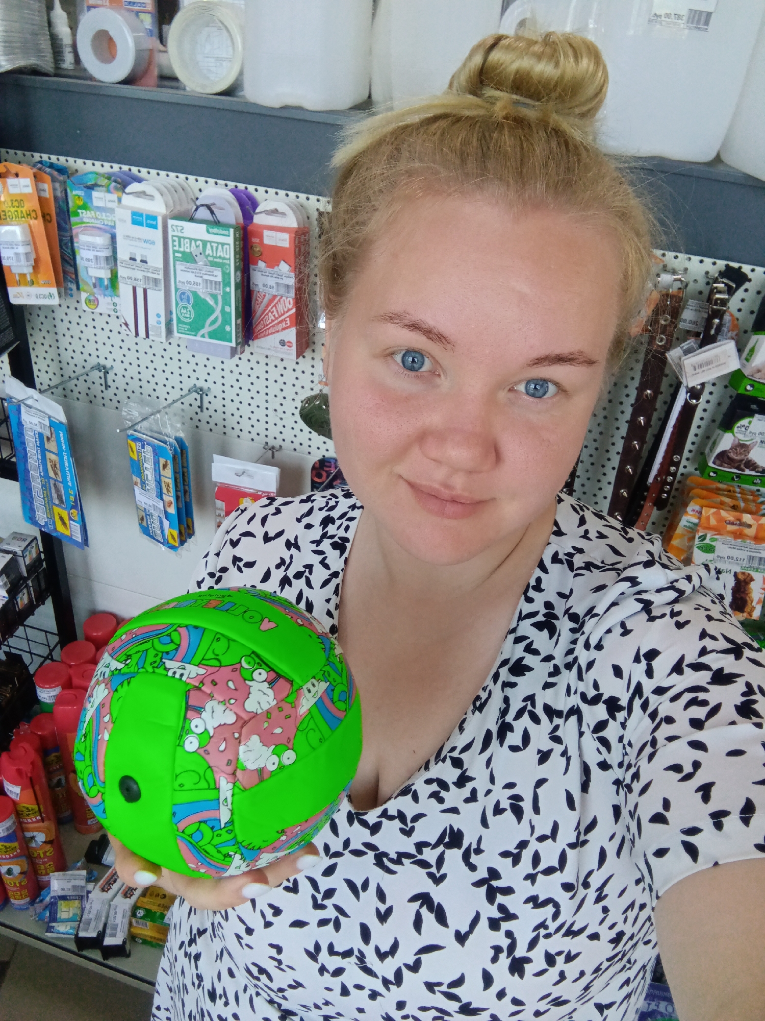 A young woman with blonde hair in a bun and blue eyes is taking a selfie in what appears to be a store. She is holding a bright green and pink patterned ball. She is wearing a white dress with a black leaf pattern. The background shows shelves stocked with various products, including what seem to be electronics accessories, pet supplies, and cleaning products. The store appears to be in the city of Fokino, Russia.