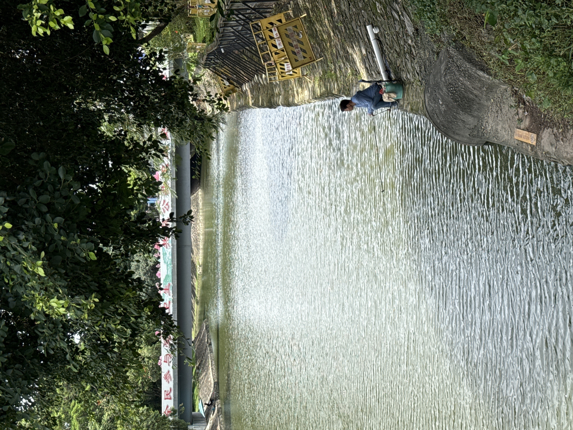 A man is fishing on the bank of a canal under a bridge. The canal's water is calm with ripples. The bank is lined with concrete and rocks. There are yellow barriers and a fence along the bank. Trees overhang the canal. In the background, text is visible on the bridge. The scene is in Tianhe District, China.