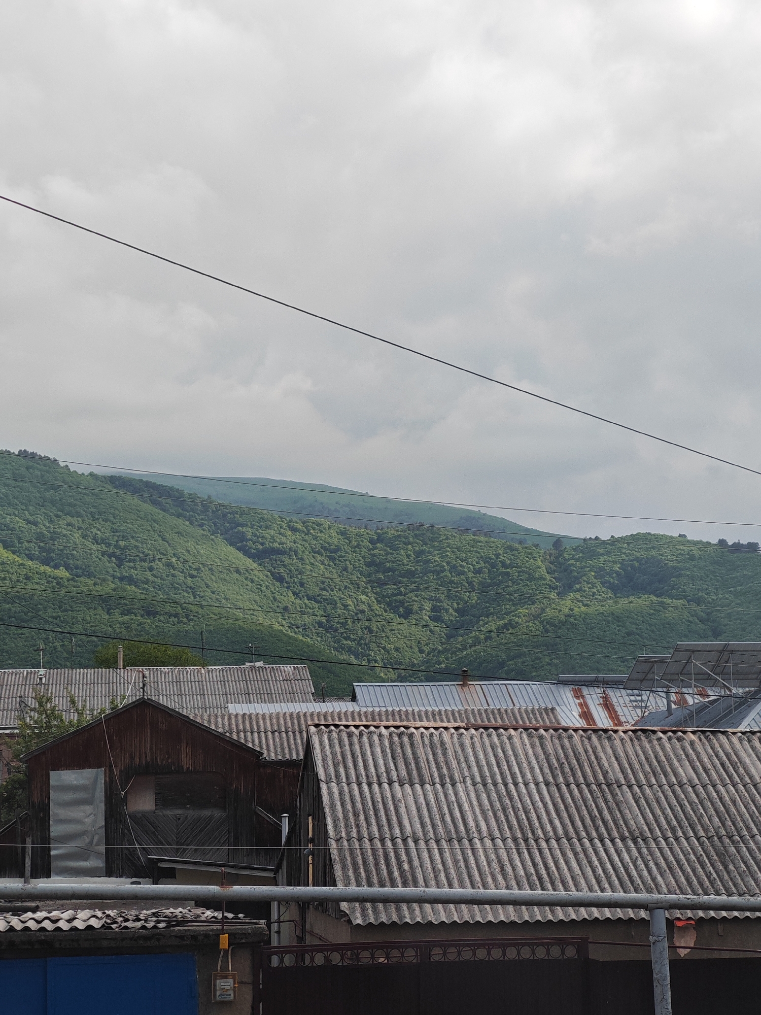 A wire runs diagonally across the frame from the upper left to the lower right, obscuring a view of a cloud-covered sky. Below the sky, a lush green mountainside is visible, covered in dense foliage. In the foreground, the roofs of several buildings are seen, some made of corrugated metal and others appearing to be slate. One wooden structure with a metal-covered door stands prominently. Power lines are visible crisscrossing the scene, and solar panels can be seen on some of the roofs. The overall impression is of a residential area nestled at the foot of a verdant mountain range. The image appears to be taken in Vanadzor, Armenia, a city known for its mountainous terrain and green landscapes.