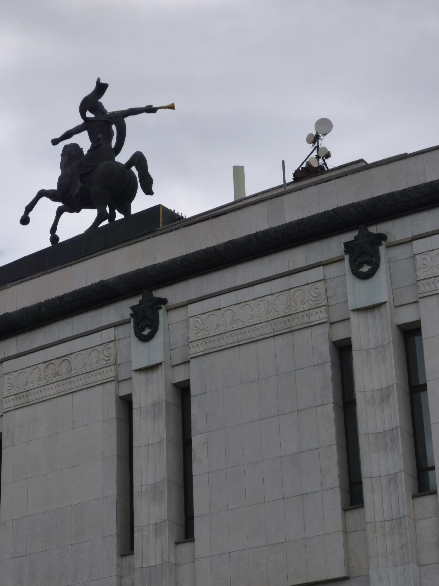 The image shows a close-up view of the building facade topped by a statue. A dark bronze statue of a person on a horse, blowing a horn, is prominently displayed against a cloudy sky. The building is constructed with large, light-colored stone blocks. Decorative elements, including a frieze with a repeating pattern of shells and a star insignia within a wreath, adorn the facade. Several tall, narrow windows are visible on the building. On the roof, several communication dishes and antennas are mounted. The overall scene suggests a historic or monumental structure, possibly in an urban setting like Moscow, Russia, given the architectural style and the Soviet-era symbol of the star.