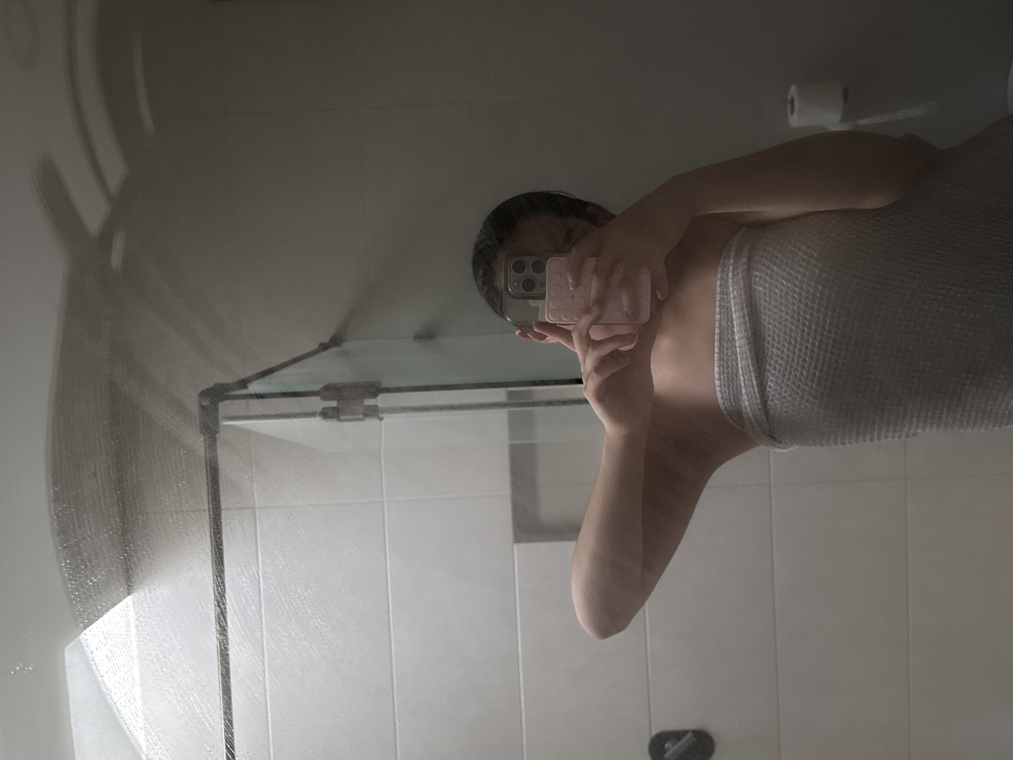 A person wrapped in a grey towel is taking a selfie in a bathroom mirror. The person is holding a pink mobile phone with a triple camera setup. The bathroom has white tiled walls and a glass shower door. A roll of toilet paper is mounted on the wall. No information is provided in the image to support the location being in Puebla, Mexico.