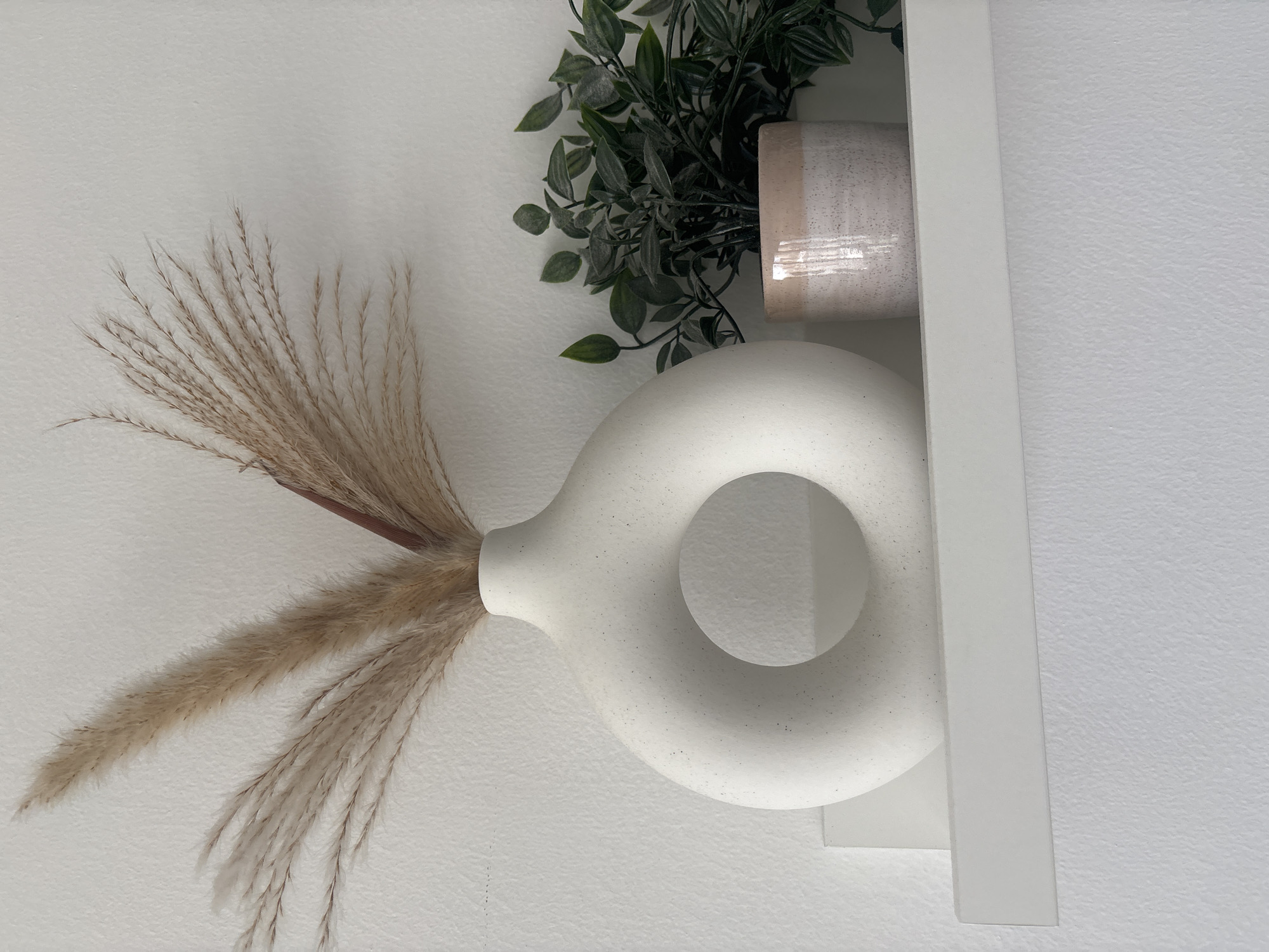 A close-up, eye-level, vertical shot captures a minimalist decor arrangement against a white, textured wall. A large, white, doughnut-shaped vase with a speckled finish stands prominently on a white shelf. Inside the vase are dried ornamental grasses with feathery plumes in a light brown hue. To the right of the vase, a small, pink and white speckled ceramic pot holds a faux green plant with multiple leaves. The shelf appears to be a floating shelf mounted on the wall. The lighting is soft and natural, casting subtle shadows. There are no people or animals present. The scene depicts a clean, modern interior aesthetic.