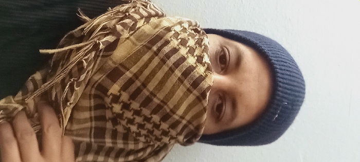 A person wearing a dark blue beanie and a brown and white checkered scarf that covers the lower half of their face. Only their eyes are visible above the scarf. The person's hand is visible on the left side of the frame, holding the scarf. The background is a plain white wall, and a dark green shirt is partially visible on the left side of the person. The image is slightly rotated counterclockwise.