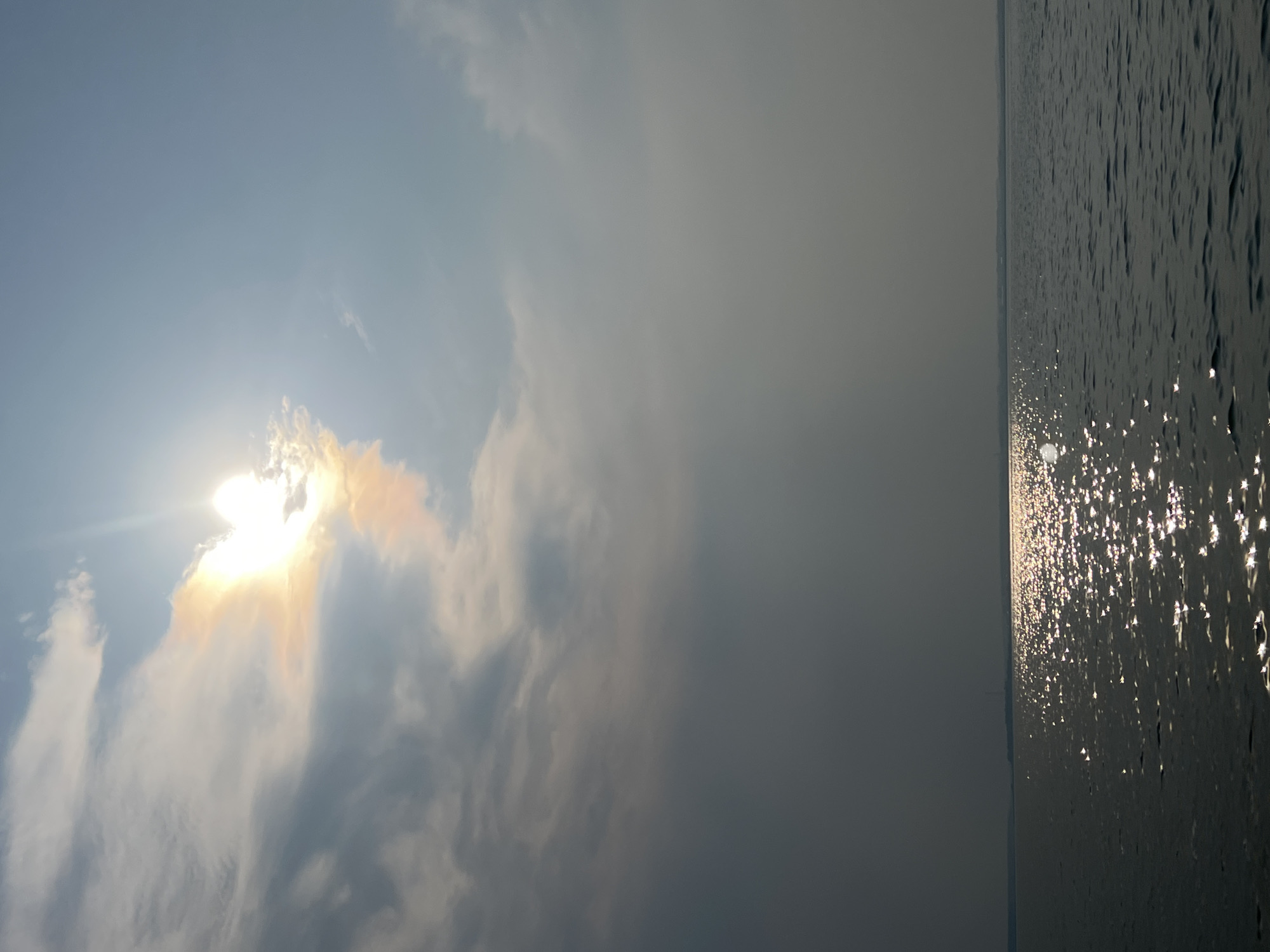 The image captures a wide expanse of water under a hazy sky with the sun partially obscured by clouds. The sun, located in the upper left quadrant, casts a bright glow, creating a rainbow effect on the surrounding clouds. The water's surface is rippled and reflects the sunlight in a multitude of sparkling points. A distant, dark landmass with faint structures is visible along the horizon. The overall atmosphere is muted due to the hazy conditions. The image does not contain any people or identifiable structures within the city of Novopichugovo, Russia.