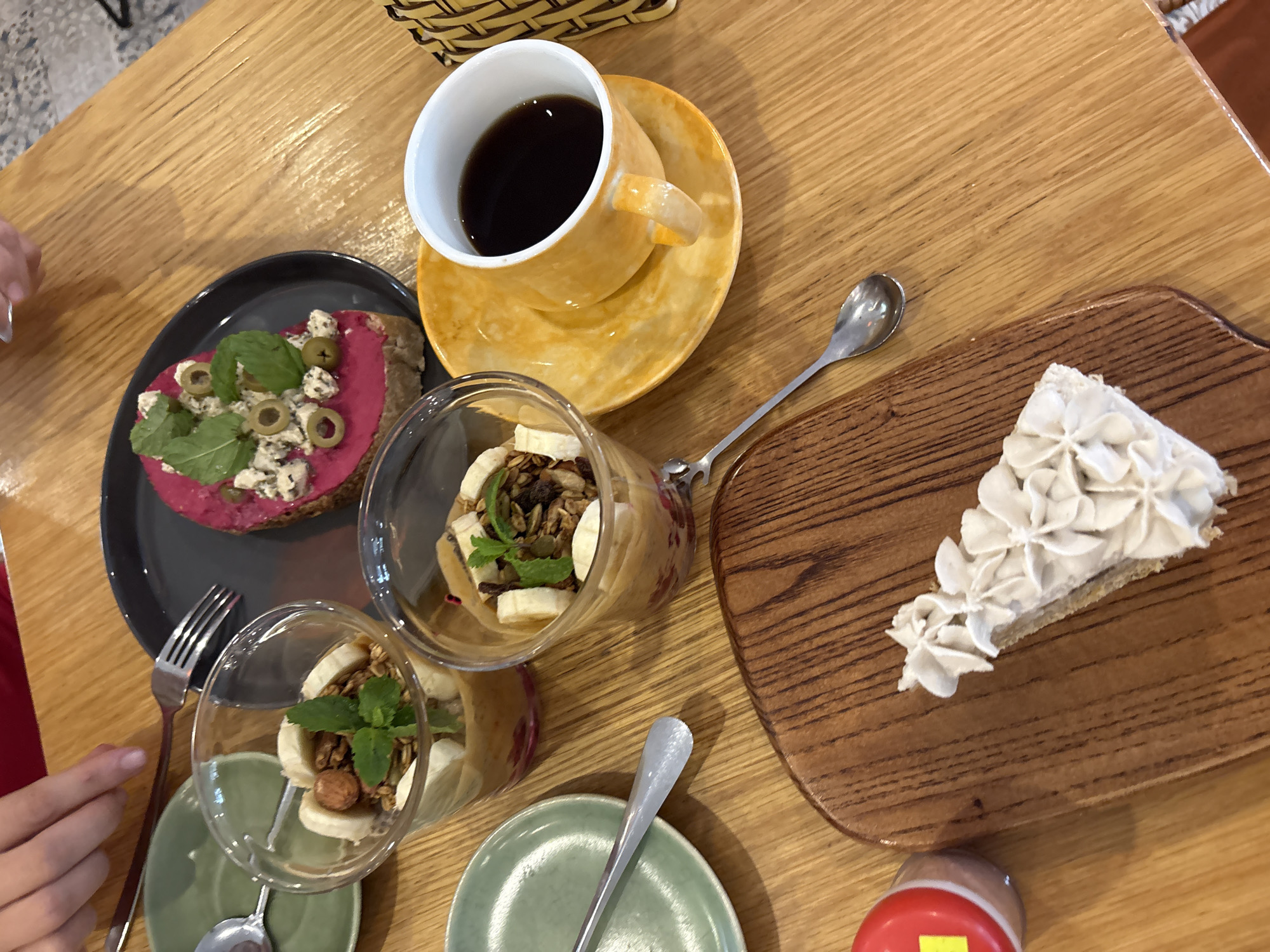 The image depicts a table setting with various food items and a cup of coffee. There is a piece of toast with a beetroot spread, topped with what appears to be cheese, olives, and a mint leaf on a grey plate. Adjacent to it, there are two small clear glasses filled with a layered dessert, possibly containing granola, bananas, and a mint leaf. A yellow cup and saucer containing dark coffee sits near the center of the table. To the right, a slice of cake with white frosting is presented on a wooden board. Silverware is placed near the food items. The scene is set on a wooden table. The location is Đà Nẵng, Vietnam.