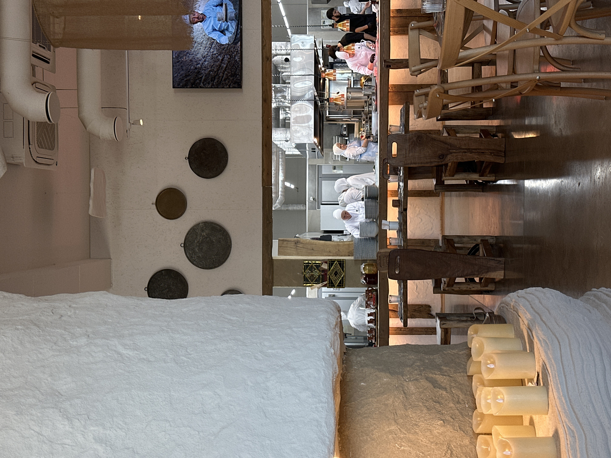The image captures the interior of a restaurant, showcasing an open kitchen, a dining area, and distinct decorative elements. The perspective is from a dining space looking towards the kitchen and another seating area.
In the brightly lit open kitchen, several chefs in white or light-colored uniforms are visible, engaged in various tasks. The kitchen features modern stainless steel surfaces and equipment. Above the kitchen pass, there are warm-toned pendant lights.
The dining areas are characterized by a blend of rustic and contemporary design. In the foreground, a prominent, light-colored, heavily textured surface dominates the lower left, resembling a large decorative wall or counter. To its right, a similar textured surface holds a cluster of glowing faux candles, casting a warm ambiance. Adjacent to this, numerous wooden chairs and tables, some stacked or arranged, suggest a seating area with a rustic aesthetic. The chairs exhibit a distinct, almost sculptural wooden design.
Along a dividing wall between the foreground and the kitchen, several dark, circular decorative plates or medallions are mounted. Above these, in the upper left, white industrial-style pipes or ventilation ducts run along the ceiling. A piece of coarse, woven, burlap-like fabric hangs from the ceiling in the upper left corner. A framed picture or screen on the far left wall displays an image of a person in a blue shirt and head covering, sitting amidst a textured, possibly agricultural, background.
The lighting in the restaurant creates a varied atmosphere; the kitchen is brightly illuminated, while the dining areas feature softer, warmer lighting, particularly from the candles. No specific activity beyond the chefs working is clearly depicted. No legible text is visible in the image. The overall impression is one of a stylish restaurant with a unique fusion of modern and rustic design.