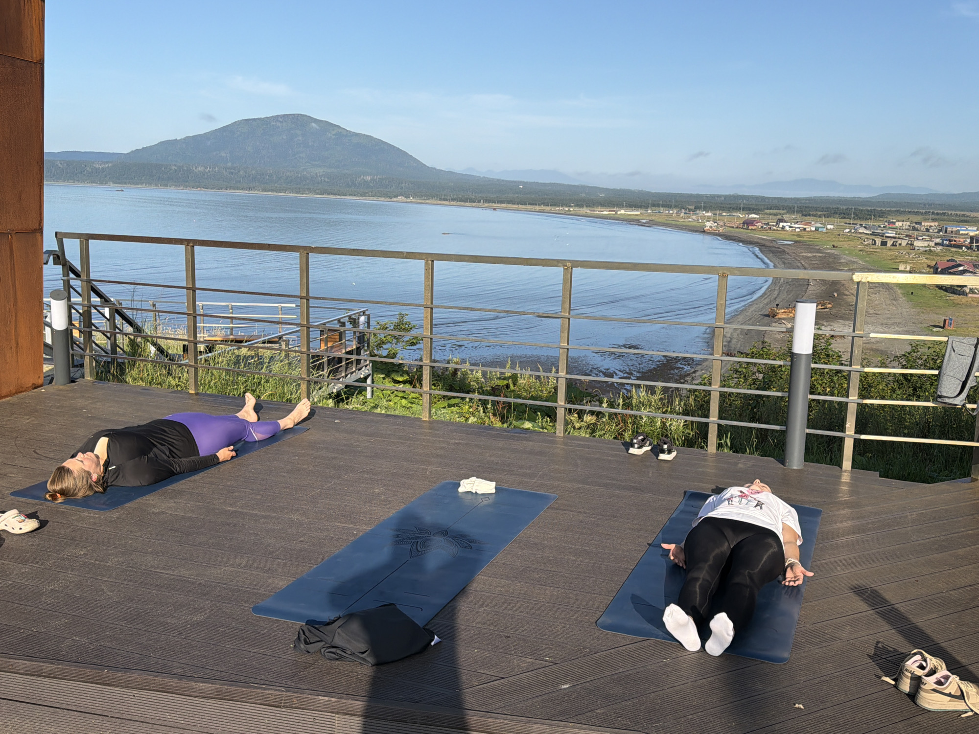 Two individuals are practicing yoga on a wooden deck overlooking a bay with a distant mountain range and a settlement. The person on the left is lying on their back on a dark blue yoga mat, wearing a black long-sleeved top and purple leggings. To their left, a white Croc shoe is visible. The person on the right is also lying on their back on a dark blue yoga mat, dressed in a white t-shirt and black leggings, with white socks on their feet. Adjacent to their mat, a black garment is placed. Several pairs of athletic shoes are scattered on the deck. A metal railing separates the deck from a grassy area and the water. The environment suggests a peaceful outdoor setting with a clear sky and calm water. The location is identified as Yuzhno-Kurilsk, Russia.