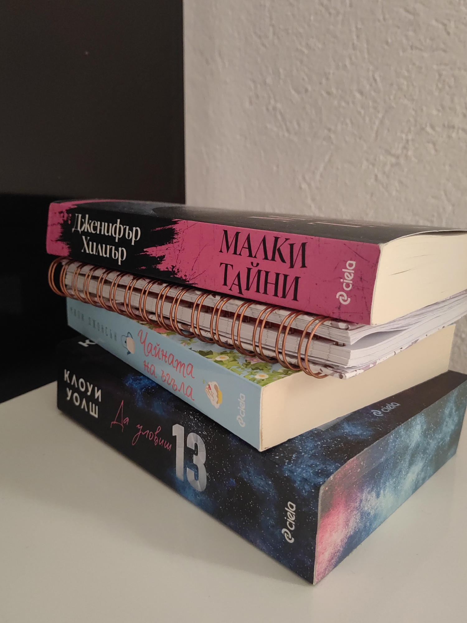 A stack of four books rests on a white surface, with a dark, vertical object to the left and a textured, off-white wall in the background. The top book has a black cover with pink text that reads "Дженифър Хипър" (Jennifer Hippen) and "МАЛКИ ТАЙНИ" (Small Secrets). The second item is a spiral-bound notebook with a copper-colored spiral. The third item, a book with a light blue cover, features floral illustrations and text that reads "Чайката на въгла" (The Seagull on the Pillow). The bottom book has a dark blue cover with a galaxy-like pattern and pink accents, displaying the text "КЛОУИ УОЛШ" (Chloe Walsh) and "Да уловиш 13" (To Catch 13). The logo "@ciela" is visible on several of the books. The lighting suggests an indoor setting, likely a room. No people or animals are present. The image is taken from a low angle, looking up at the stack of books. The scene is static and there are no discernible actions taking place. The specific location of Djerovo, Bulgaria cannot be determined from the visual information provided.
