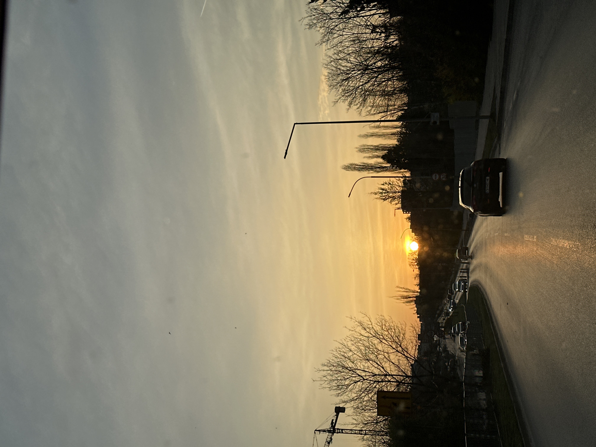 The image shows a sunset view from a vehicle on a road in Sofia, Bulgaria. The sky is a mix of gray and orange hues, with the sun setting between trees and buildings. A black car is driving on the right side of the road, and other vehicles are visible further ahead. Streetlights are present along the roadside. In the lower left, a construction crane is visible. The road surface appears wet and reflects the sunlight.