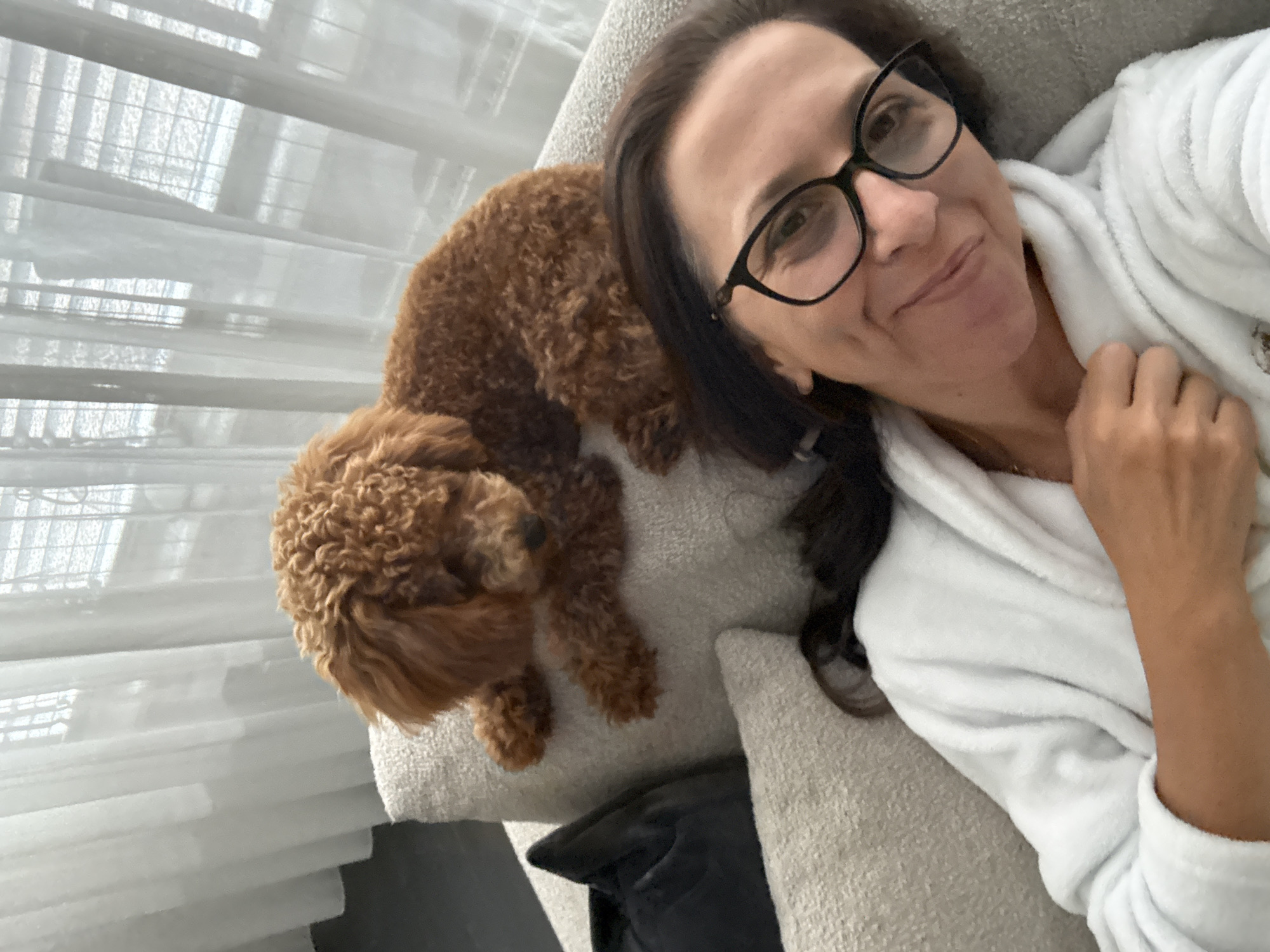 A brown poodle is perched on the back of a couch behind a woman wearing glasses and a white bathrobe. The woman is taking a selfie, smiling at the camera. The background shows sheer white curtains and blinds. The image appears to be taken indoors, likely in a residential setting.