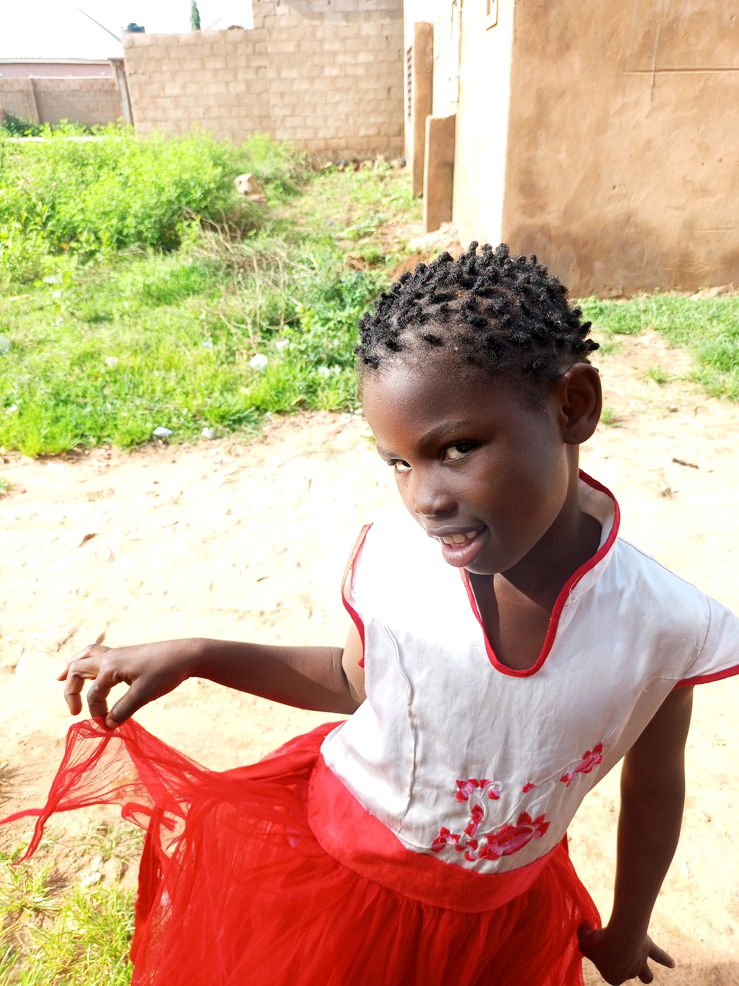 A young Black girl with short, braided hair and dark skin is standing outside in what appears to be a rural or undeveloped area, possibly in Tempure, Nigeria. She is wearing a white shirt with red floral embroidery and a bright red skirt. Her left hand is raised, holding the edge of her skirt, while her right arm hangs by her side. She is looking towards the camera with an open mouth and a slight smile. Behind her, there is a sandy ground with patches of green grass and some debris. In the background, there are unfinished brick buildings and more vegetation. The lighting suggests it is daytime, with bright sunlight casting shadows.
