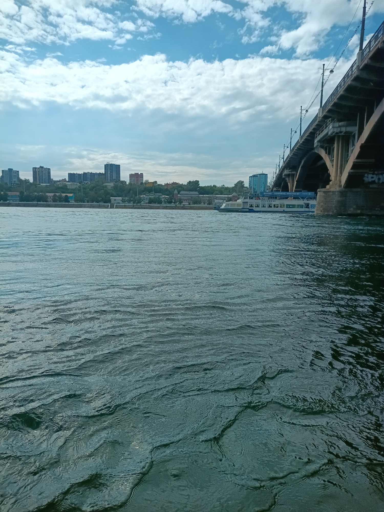 The image depicts a wide river with moderate waves under a partly cloudy sky. On the right side of the frame, a large concrete bridge arches over the water. The bridge has multiple supporting pillars and a roadway on top. A white and blue passenger boat is visible on the river, passing under the bridge. In the background, a cityscape with several high-rise buildings and trees lines the riverbank. The overall scene suggests an urban environment in Russia, likely Irkutsk, given the presence of a significant river and bridge infrastructure typical of cities in that region. No people are clearly visible in the image.