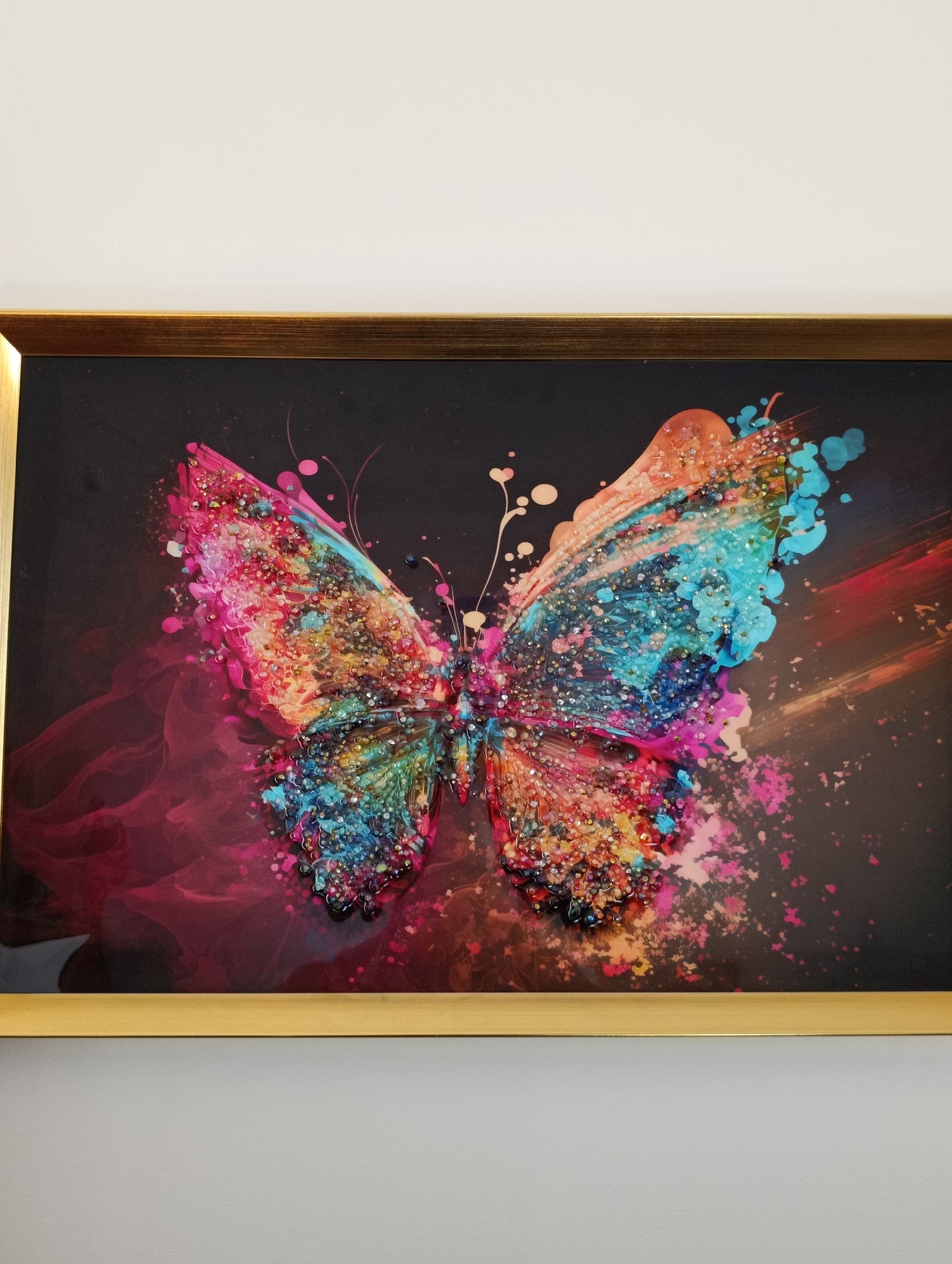 The image displays a framed artwork featuring a vibrant, abstract butterfly. The butterfly's wings are a kaleidoscope of colors including pink, orange, blue, and teal, adorned with what appears to be glitter or small beads that add a textured, shimmering effect. The background is dark, with swirling wisps of pink and red smoke-like patterns and scattered flecks of color. The artwork is housed in a thin, golden frame and is mounted on a plain, light-colored wall. No specific location in Setúbal, Portugal, can be identified from the image.