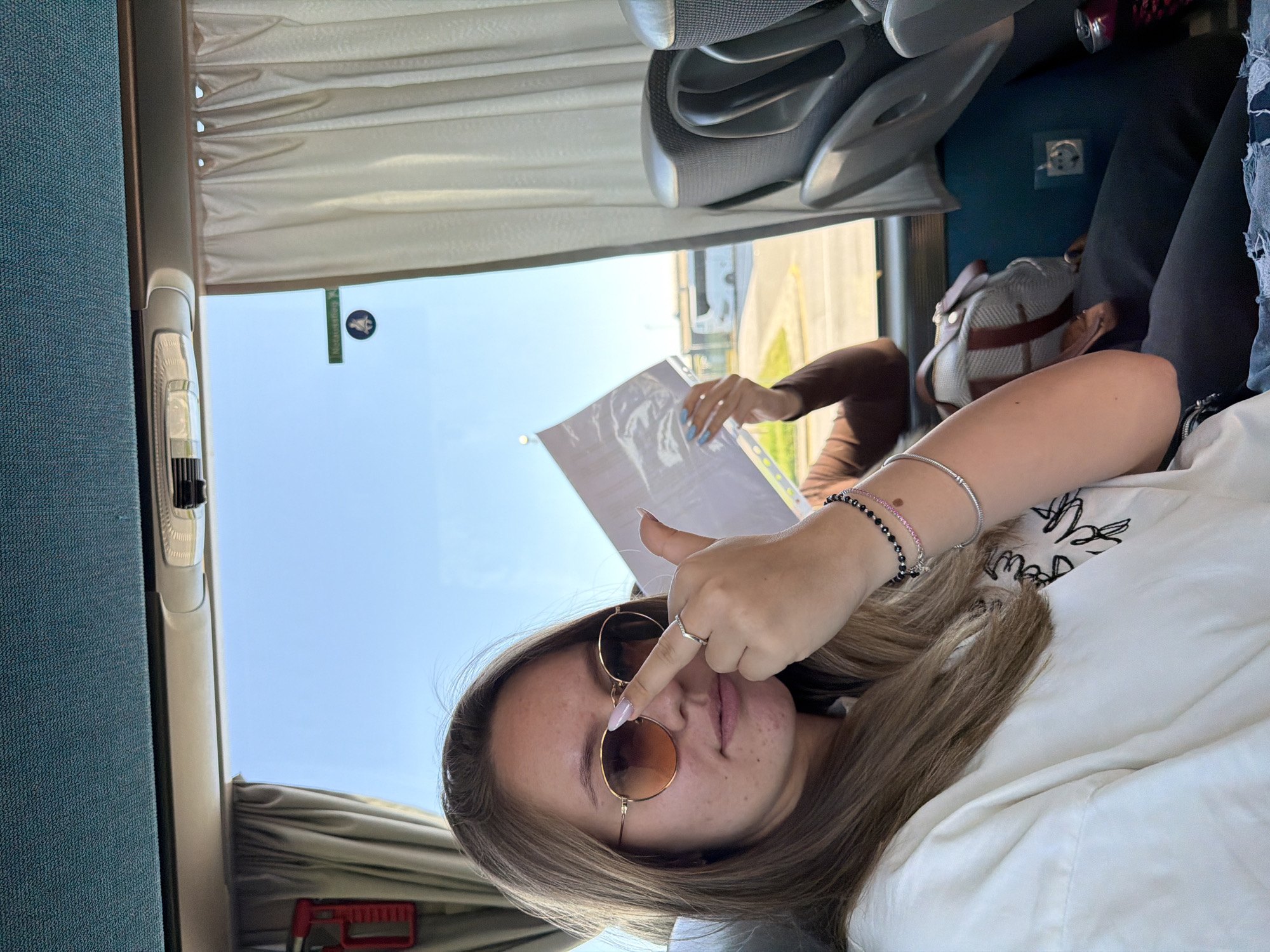 A selfie taken inside a bus in Kapitan Andreevo, Bulgaria. A young woman wearing round sunglasses and multiple bracelets is pointing at the lens with her index finger while holding a document. The background shows a bus window, curtains, and seats. A person in a brown shirt is visible partially in the window.