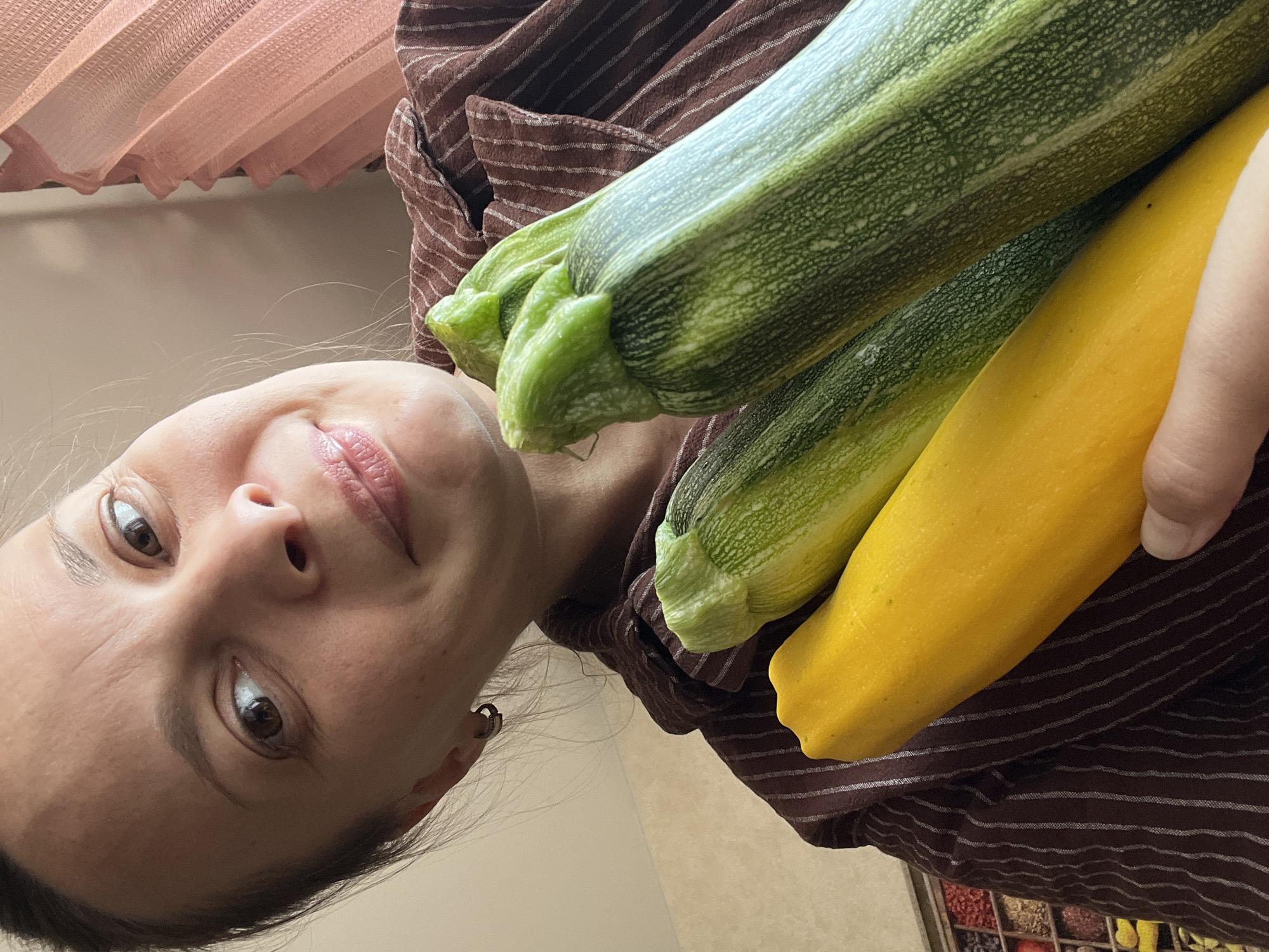 The media file is a vertically oriented, eye-level, close-up photo of a woman holding three zucchinis. She is looking at the camera with a slight smile, her face taking up the left side of the frame. She has dark hair, brown eyes, and is wearing a brown striped shirt. The zucchinis, two green and one yellow, are positioned in the upper right portion of the frame, held by her hand, which is visible at the far right edge. The background appears to be an indoor setting, with a blurred pink curtain in the upper left corner and a beige wall or surface behind the woman and the vegetables. The lighting suggests it is daytime, likely indoors. The image is a selfie, indicated by the angle and the woman looking directly at the camera. The overall mood is casual and possibly reflects a moment of showcasing fresh produce. No visible text is present in the image.