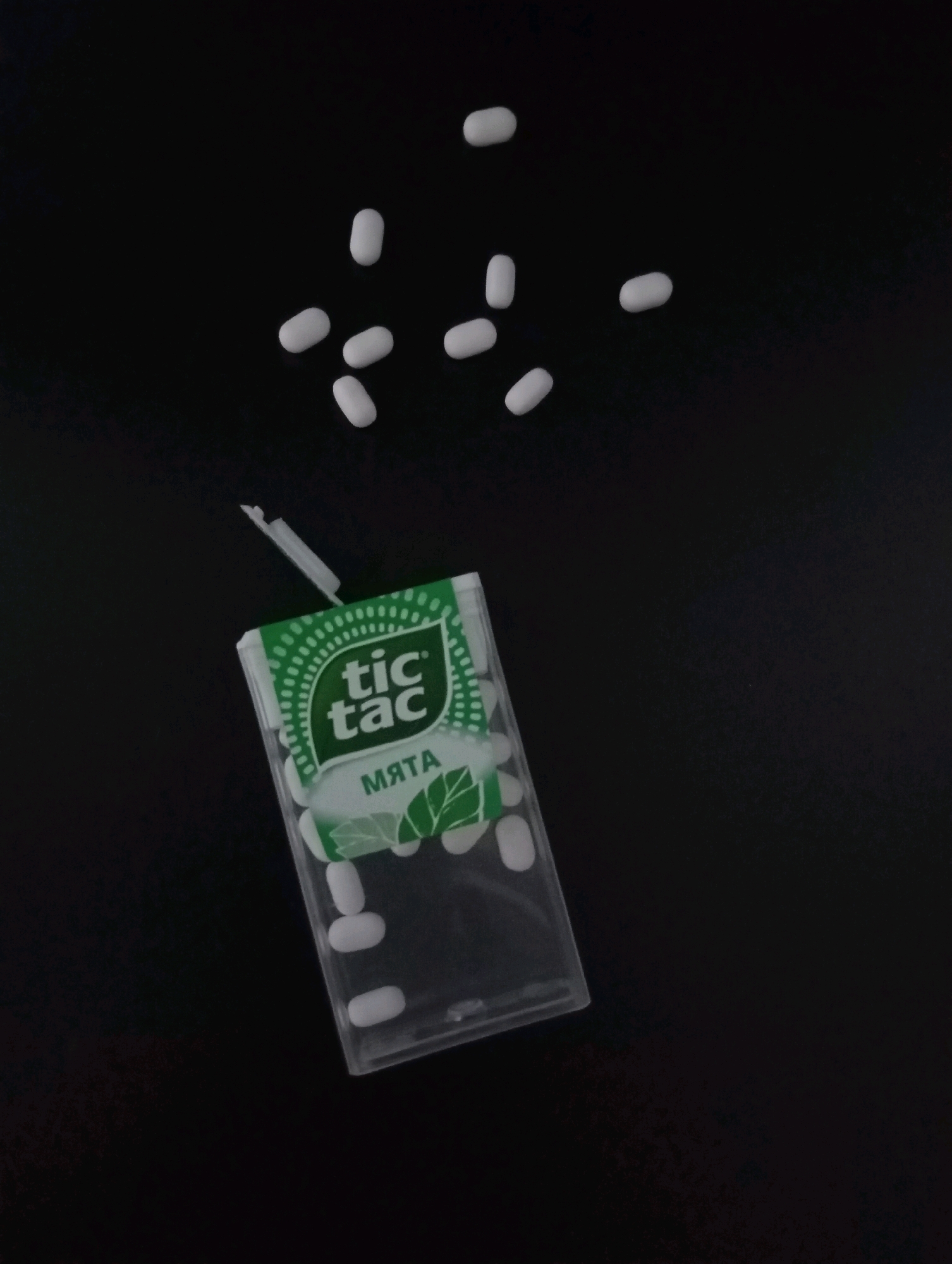 The image shows a Tic Tac mint dispenser and several loose mints scattered on a black surface. The dispenser is partially open, revealing white mints inside. Some mints are also visible outside the dispenser and scattered in the upper right portion of the image. The background is a plain black surface, suggesting an indoor setting. There are no people or indications of a specific location in Krasnodar, Russia, present in the image.