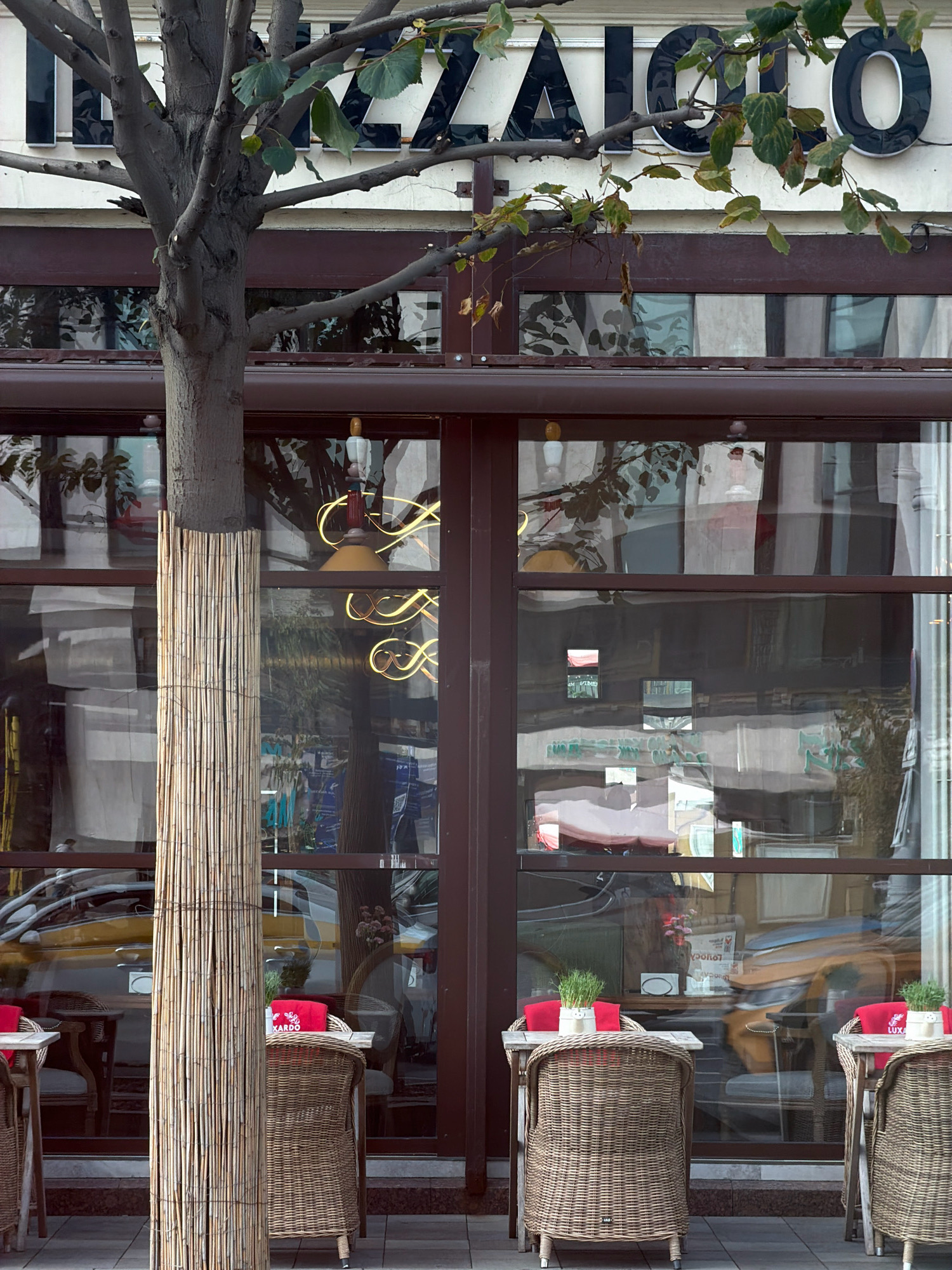 The image captures a street-level view of a cafe's exterior in Moscow, Russia. A large tree with bare branches partially obscures the building's signage, which features dark, stylized letters spelling out "IAIO". The cafe has large, dark-framed windows that reflect the surrounding environment, including other buildings, vehicles, and street scenes.
In the foreground, several tables are set up outside the cafe. Two tables are made of light-colored wood with wicker chairs surrounding them. One table has a red napkin and a small white pot with green grass. The other table, partially visible on the left, also appears to have a red element. A third table, also with a wicker chair, is seen further to the right, with a red napkin displaying the word "LUXA" and a small white pot of greenery.
A prominent element in the foreground is a thick tree trunk wrapped in a tall, vertical covering of bundled reeds or bamboo stalks, secured with twine. The ground in front of the cafe is paved with light-colored tiles.
The reflections in the cafe windows are complex and layered, showing glimpses of yellow vehicles, indoor lighting fixtures with yellow spiral designs, and other interior details that suggest a seating area with white curtains. There are also abstract reflections of buildings and sky. The overall impression is a European-style cafe setting, likely in an urban area of Moscow.