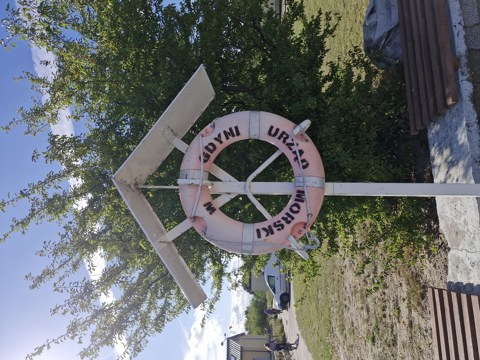 The image shows a pink life preserver with black lettering that reads "URZĄD MORSKI GDYNI" in a circular pattern. The life preserver is mounted on a white pole with a white, angled roof-like structure above it. Behind the life preserver is a lush green tree. In the background, there are two people on bicycles riding on a paved path. To the left of the path is a light-colored building with a grey roof and windows. Further in the background, a white van is parked. The ground is a mix of grass and dry soil. The sky is blue with scattered white clouds, and the sun is shining brightly, casting shadows. There is a wooden bench in the lower right corner of the image. The scene is outdoors, suggesting a public area, possibly near the sea given the presence of the life preserver and the inscription referring to a Maritime Office. The location is identified as Jastarnia, Poland.
