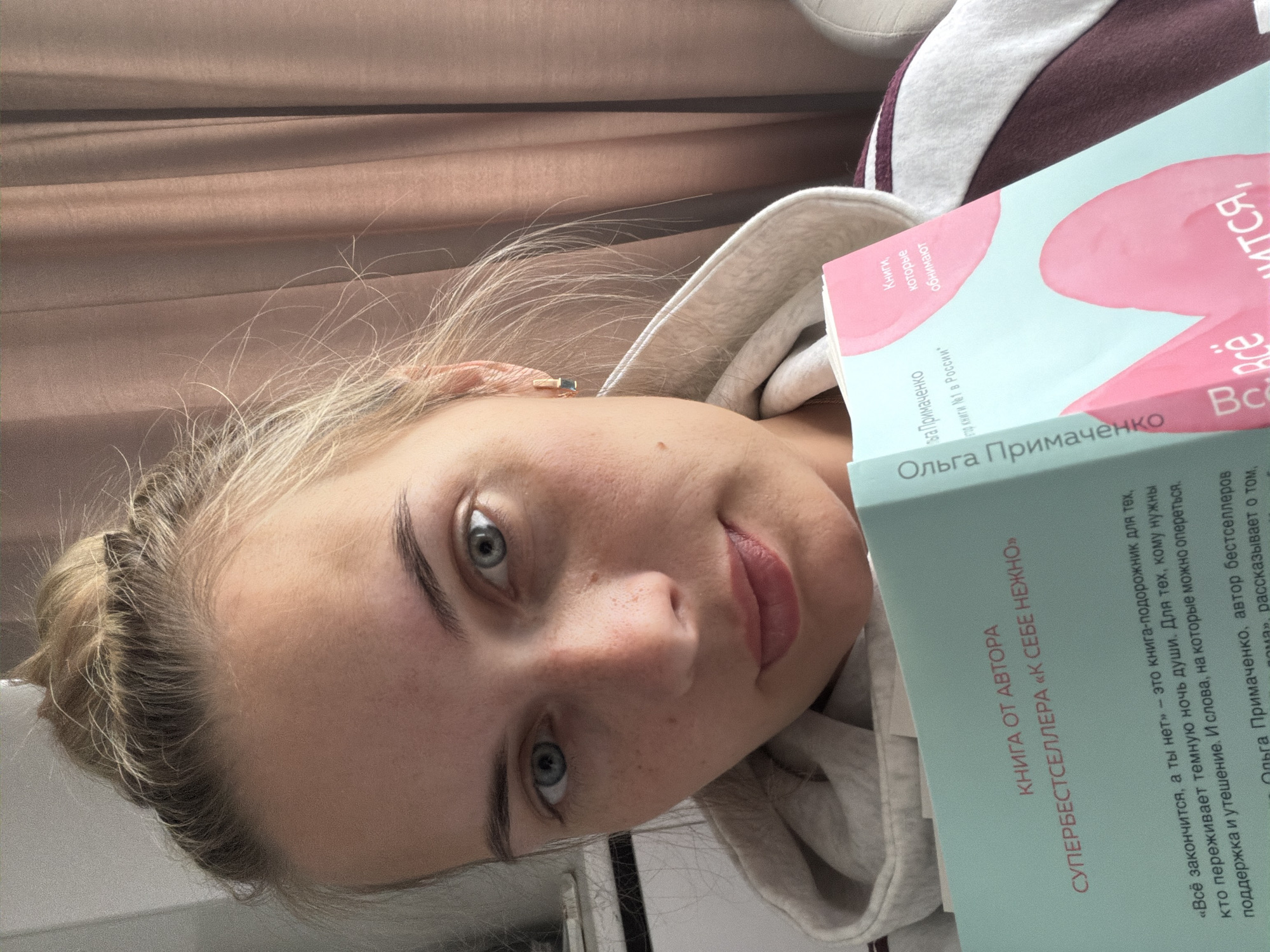 A young woman with blonde hair pulled back in a bun, wearing a grey hooded sweatshirt with maroon trim, is holding two books. The book closest to the viewer has a light blue cover with the title "Ольга Примаченко" (Olga Primachenko) written in Cyrillic. Beneath the title are the words "КНИГА ОТ АВТОРА" (BOOK FROM THE AUTHOR) and "СУПЕРБЕСТСЕЛЛЕРА «К СЕБЕ НЕЖНО»" (SUPERBESTSELLER "TO YOURSELF GENTLY"). The second book has a pink and light blue cover with Cyrillic text. The text on the back of the book is visible and is written in Russian.
The background consists of a brown draped cloth.