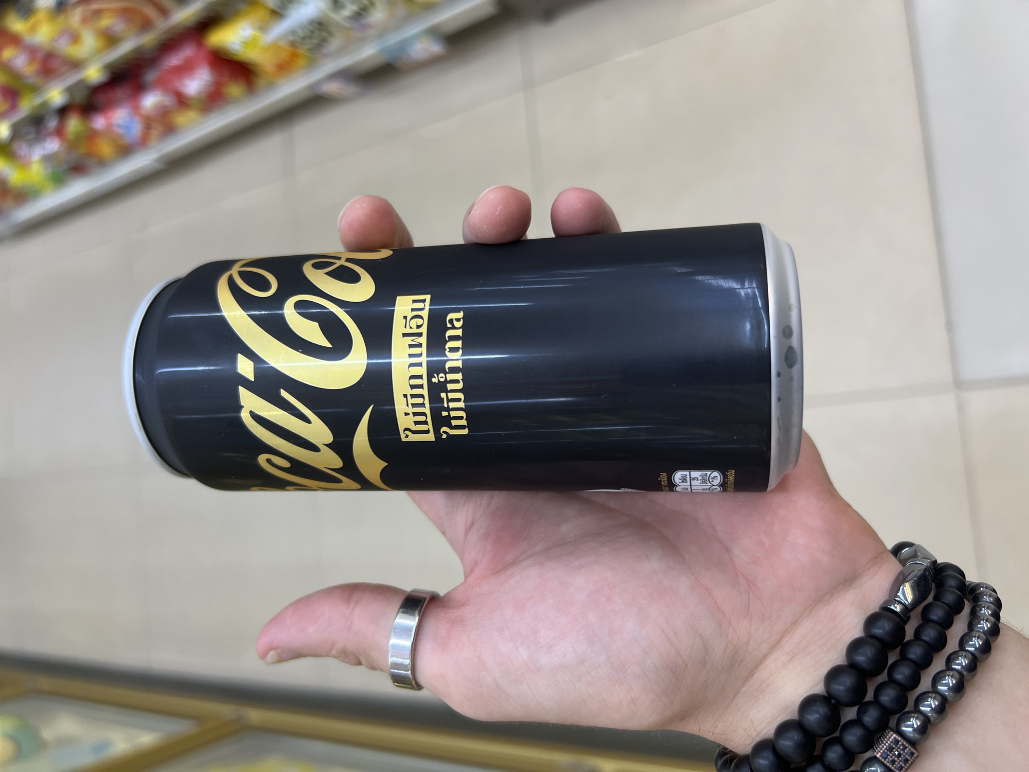 In Choeng Thale, Thailand, a person's hand is holding a black Coca-Cola can with gold lettering. The can is in the foreground, with the Coca-Cola logo prominently displayed in gold. Below the logo, there is text in Thai. The person is wearing a silver ring on their index finger and a bracelet made of dark beads on their wrist. The background shows blurred shelves with various products, suggesting the can was purchased or is being held in a retail setting like a convenience store or supermarket. The lighting appears to be artificial, consistent with an indoor environment.