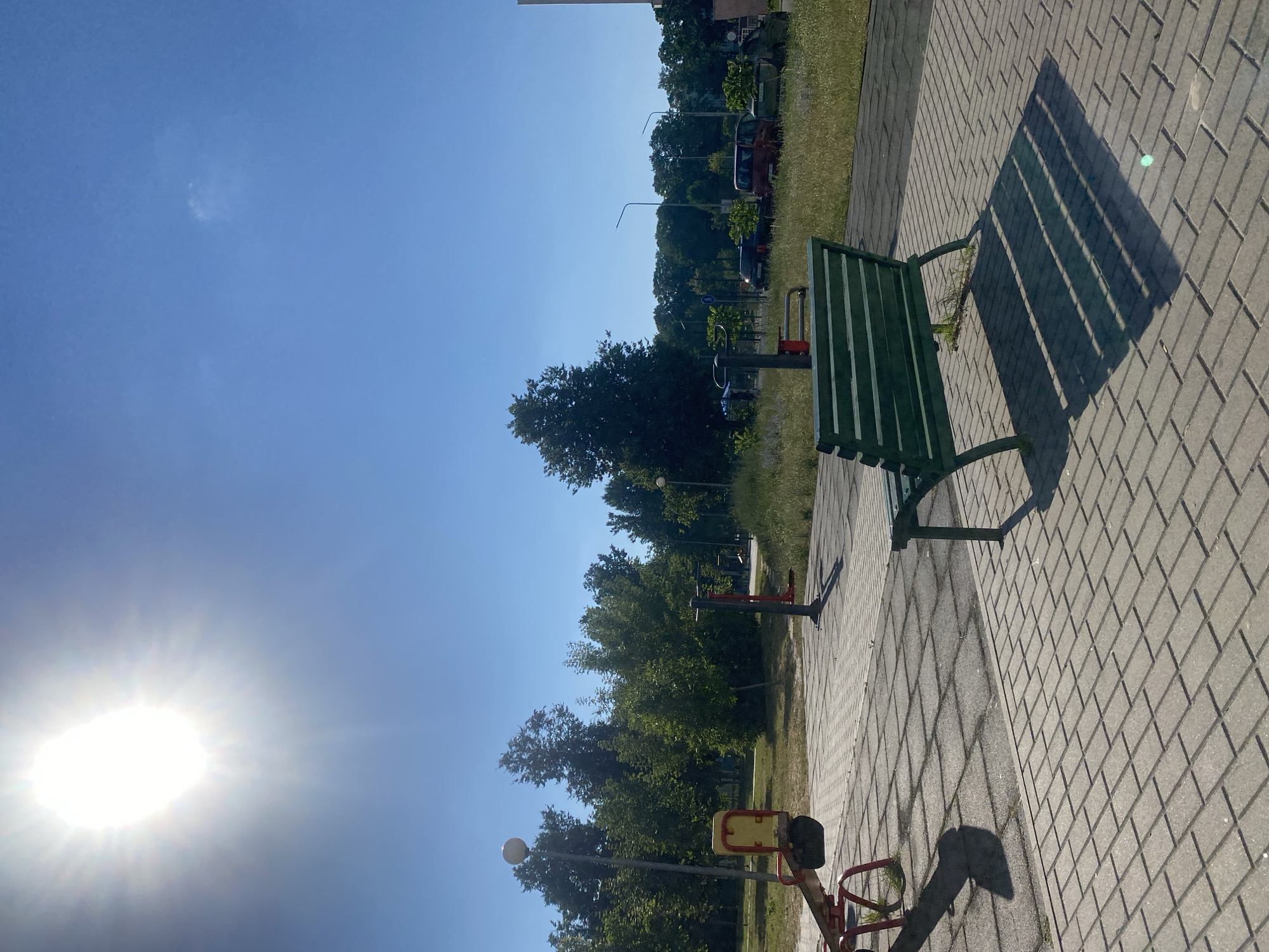 The image depicts an outdoor scene with a bright, sunny sky. The sun, shining intensely and with visible rays, dominates the left side of the frame. A green metal bench is positioned on a paved walkway made of rectangular grey bricks. The bench casts a long, striped shadow on the walkway, indicating it is likely midday or early afternoon.
Beyond the walkway, a grassy area is visible, bordered by a line of trees. Further back, there are some buildings, streetlights, and parked cars, suggesting an urban or suburban setting. Several pieces of outdoor exercise equipment, painted in red and yellow, are also present in the grassy area. One piece, in the foreground, appears to be a seesaw-like apparatus.
The overall mood is peaceful and bright, suggesting a pleasant day for outdoor activity. There is no visible text in the image. The location context provided is Homyel, Belarus.
