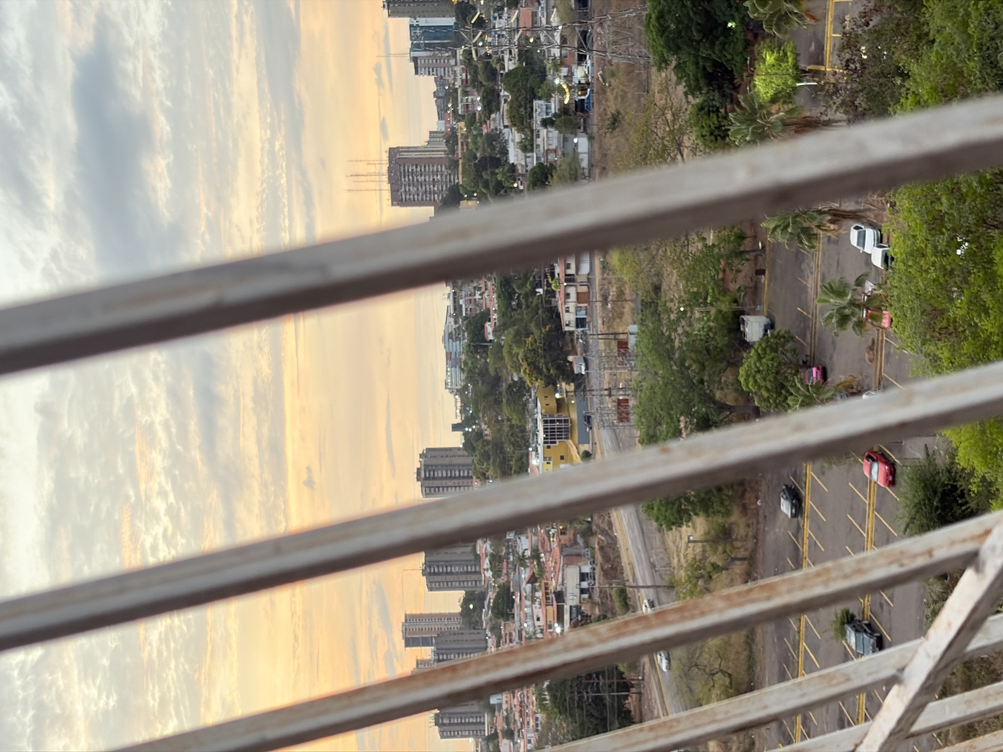 The image, taken from an elevated perspective through a metal railing, shows a cityscape at sunset. The city includes tall apartment buildings interspersed with smaller structures and trees. A parking lot with several parked cars and trees are located to the right. The sky is partially visible with clouds illuminated by the setting sun.
The image was taken from the Orinokia Mall in Ciudad Guayana, Venezuela. The mall is on a hill overlooking the city. The parking lot in the image is part of the mall. The buildings visible in the distance are residential towers.