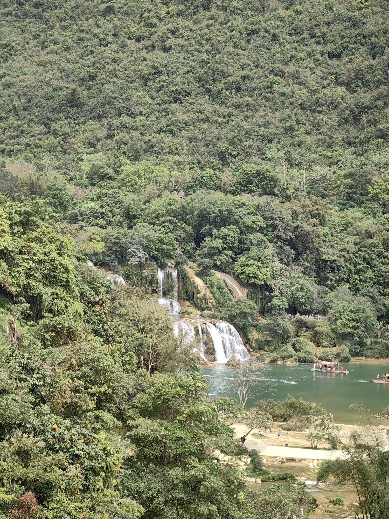 The image shows Ban Gioc Falls in Daxin County, China. The falls cascade down a rocky slope into a turquoise river. Lush green vegetation covers the surrounding hills and banks. Several boats carrying people are on the river. A small sand area is visible along the bank, with a few people walking. The environment is dense with trees and foliage.