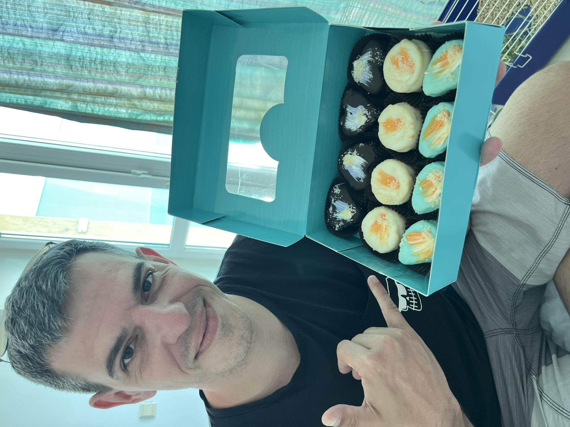 A man with short, graying hair, wearing glasses perched on his head and a black t-shirt, smiles at the camera while holding an open turquoise box of chocolates. The chocolates are arranged in two rows of six and have various toppings, including dark chocolate with white and purple drizzle, white chocolate with orange accents, and aqua-colored chocolate with orange accents. He is wearing plaid shorts and has his left hand extended, making a shaka sign. The background shows a window with vertical blinds and what appears to be a view of a building. The scene is indoors.