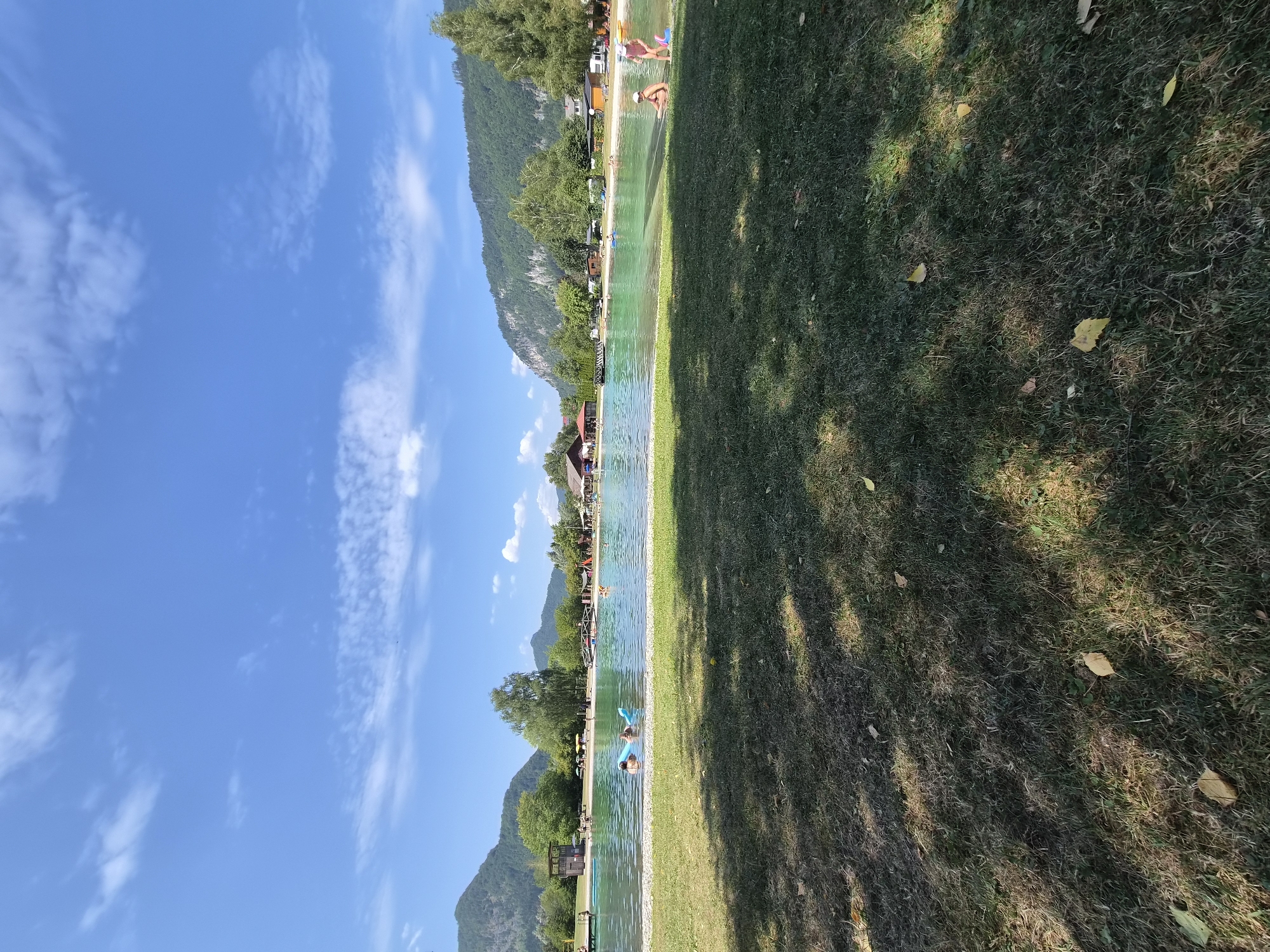 The image depicts a sunny day at a lakeside swimming area, likely the Badesee in the town of Landl, Austria, given the characteristic mountainous backdrop and lush greenery. In the foreground, a patch of dry grass is visible, with scattered yellow leaves. Beyond the grass, a clear, turquoise lake stretches out, bordered by a sandy beach. Several people are enjoying the water: two individuals are on a blue inflatable raft, one of whom is partially submerged, while others are swimming or wading. On the beach, two young children, one in a pink swimsuit and the other in white swimwear, are near the water's edge. Behind the lake, a line of trees and buildings, including what appears to be a restaurant or cafe with a red roof, are situated along the shore. A wooden bridge spans a section of the water. The background is dominated by rolling green hills and steep, rocky mountains, typical of the Austrian Alps, under a bright blue sky dotted with fluffy white clouds. The overall scene suggests a recreational summer day.