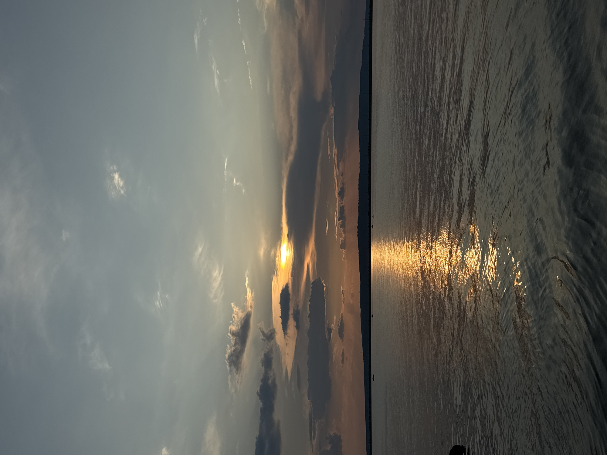 The image depicts a serene sunset over the Kama River in Chaikovsky, Russia. The sky is a gradient of soft blues and greys, with streaks of orange and yellow from the setting sun breaking through the clouds. The sun itself is partially obscured by a dark cloud, casting a golden reflection on the rippling water. The horizon is marked by a dark, silhouetted line of trees or hills. In the distance, several small boats are visible on the water, suggesting a peaceful recreational atmosphere. The overall impression is one of tranquility and natural beauty.