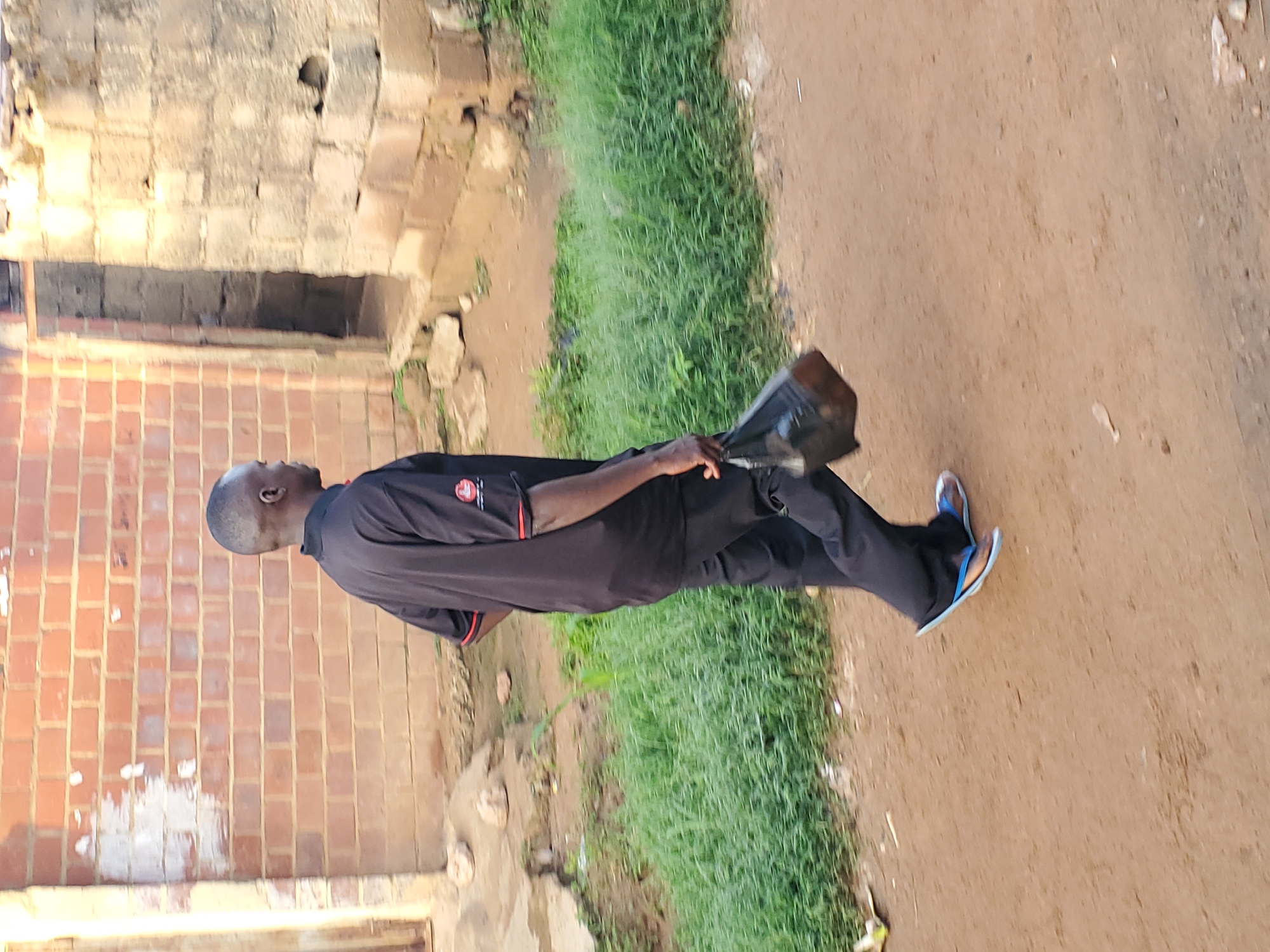 A dark-skinned man in a black shirt and pants with blue and brown flip-flops is walking to the right, carrying a black plastic bag in his right hand. He is walking on a dirt path in front of a low-lying brick building and a concrete structure. Tall green grass is to the left of the path. The man is facing away from the camera. The scene appears to be an outdoor area in the city of Jos, Nigeria.