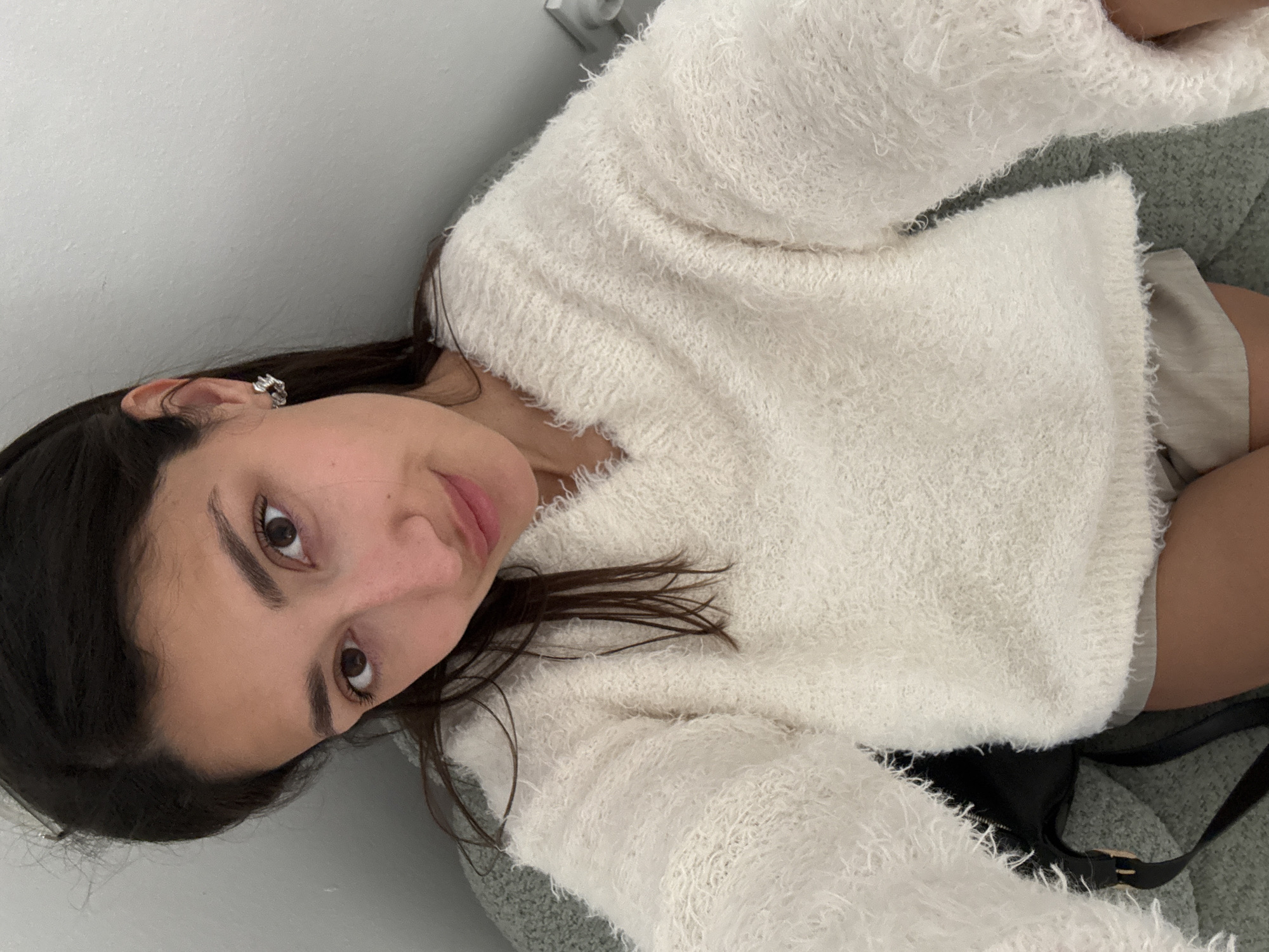 A young woman with dark hair and brown eyes is posing for a selfie. She is wearing a fluffy white sweater and an earring. She appears to be indoors, against a white wall. A portion of a purse is visible. The location is unknown.