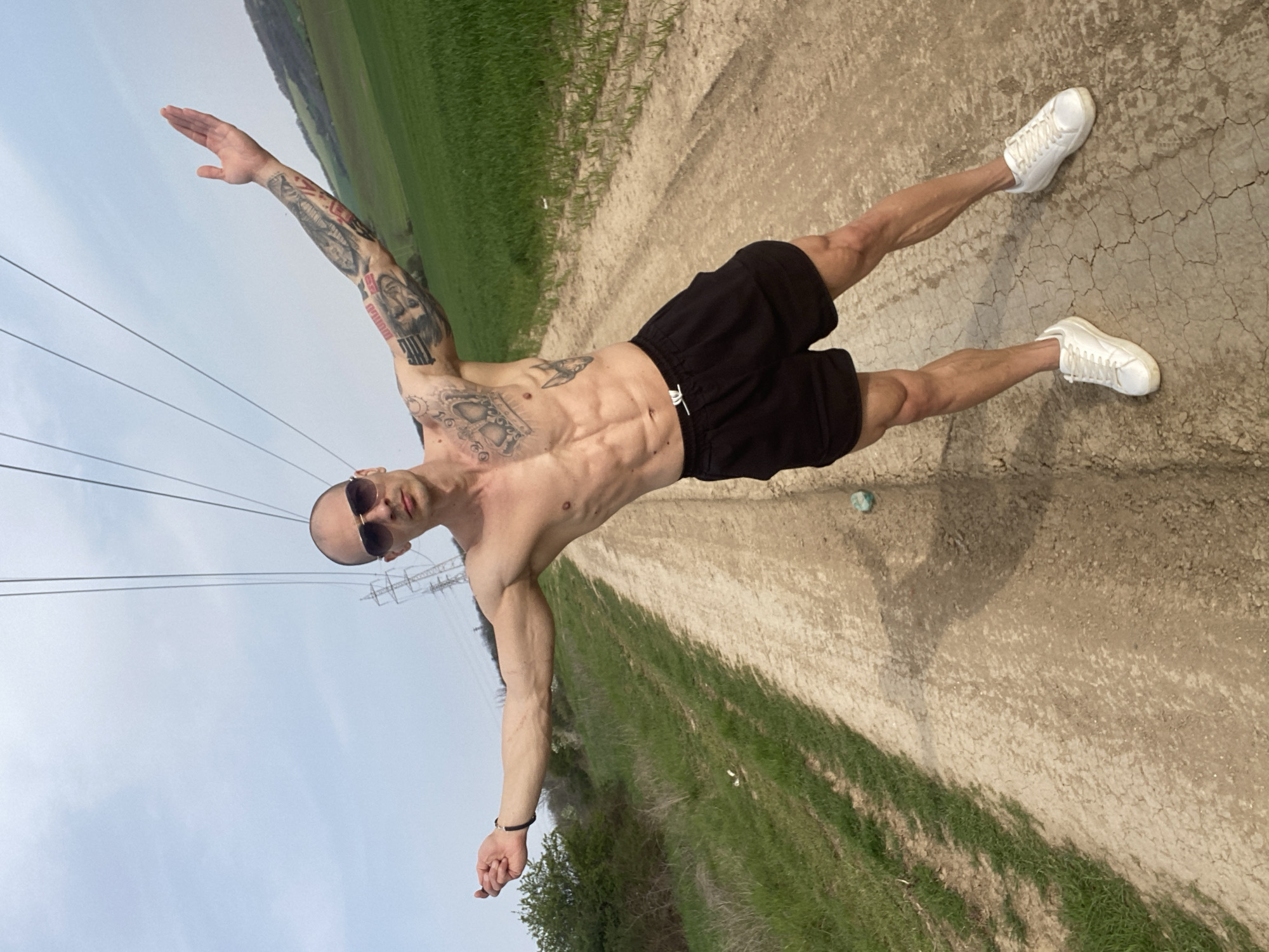 A shirtless, muscular man with a shaved head and sunglasses poses on a dirt path bordered by grassy fields in an outdoor setting. He is wearing black shorts and white sneakers. His arms are outstretched, and he has multiple tattoos on his arms and torso. Power lines stretch across the sky above him. The location appears to be near Pleven, Bulgaria, given the landscape and power line infrastructure. The dirt path is cracked and dry, with patches of green grass growing along the sides. The sky is a pale blue.