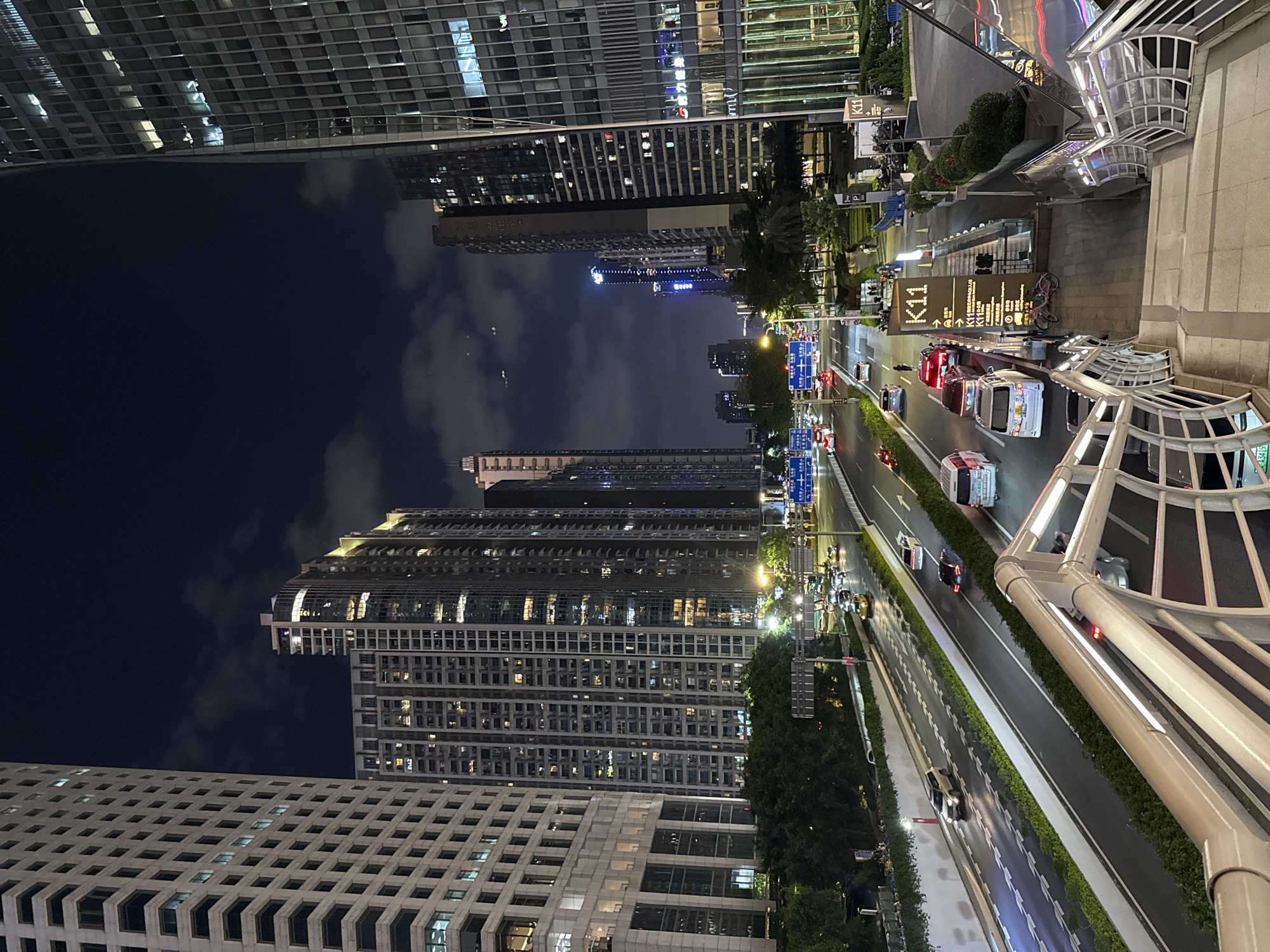 The image presents an elevated nighttime view of Tianhe District, China. Skyscrapers with illuminated windows dominate the urban landscape, contrasting with the dark sky containing scattered clouds. Below, a multi-lane road shows vehicular traffic, including cars and delivery trucks. Streetlights illuminate the scene. A signpost indicates the direction to the K11 shopping mall. A pedestrian overpass structure is visible in the foreground.
