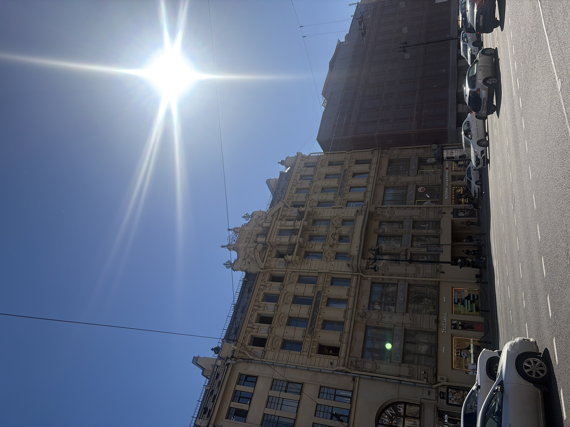The image captures a sunny day on Nevsky Prospekt in Saint Petersburg, Russia. The sun is prominently visible in the sky, creating a lens flare effect. In the foreground, a street with cars and lane markings is shown. To the left, several multi-story buildings with ornate architecture line the street. One building features the signage "Italiani" on its ground floor. To the right, another building under construction or renovation is visible, covered in dark scaffolding. Pedestrians are seen on the sidewalks in front of the buildings. The sky is clear and blue.