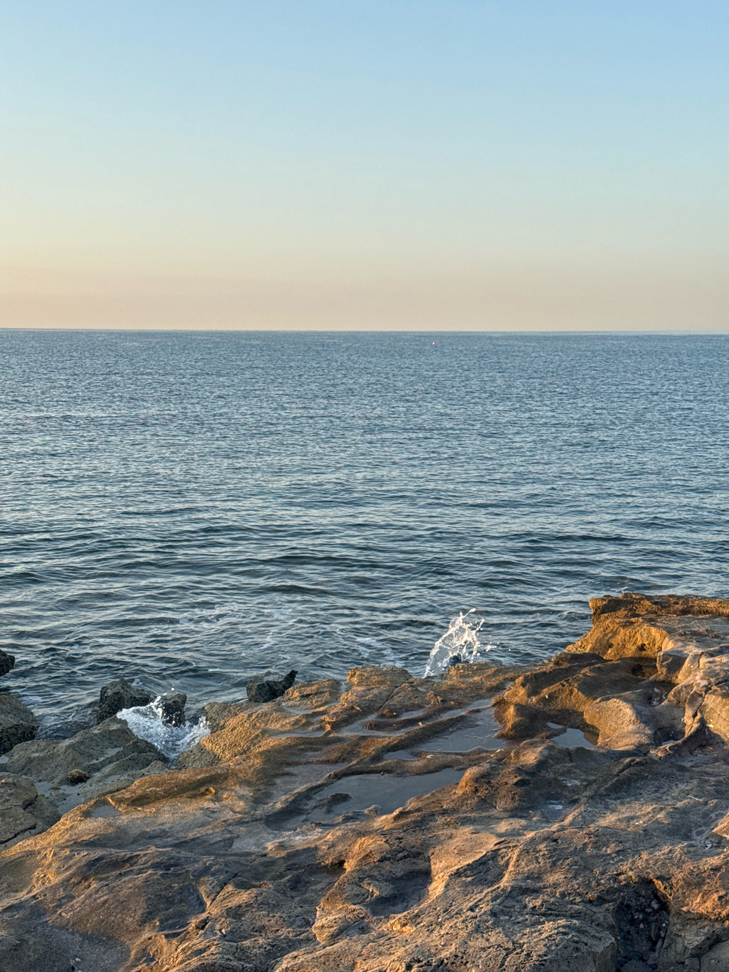 The image depicts a tranquil coastal scene in the Agios Tychona Community, Cyprus. The foreground is dominated by rough, textured rocks with pools of water reflecting the sky. Sunlight casts a warm glow on the rocks, highlighting their contours. The sea, a calm expanse of blue, stretches to the horizon under a clear, gradient sky that transitions from a pale yellow near the horizon to a light blue overhead. There are no people or visible human activity in the frame. The composition emphasizes the natural beauty of the coastline and the interplay of light on the water and rocks.