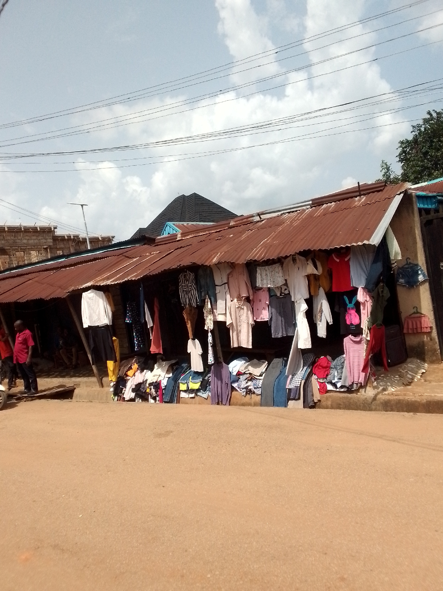A roadside second-hand clothes stall is located on a dirt road in an urban area. The stall consists of a corrugated metal roof supported by poles, with clothes hanging from the front. Some clothes are also spread out on the ground. Two people are standing to the left of the stall. In the background are buildings and trees. The sky is overcast.