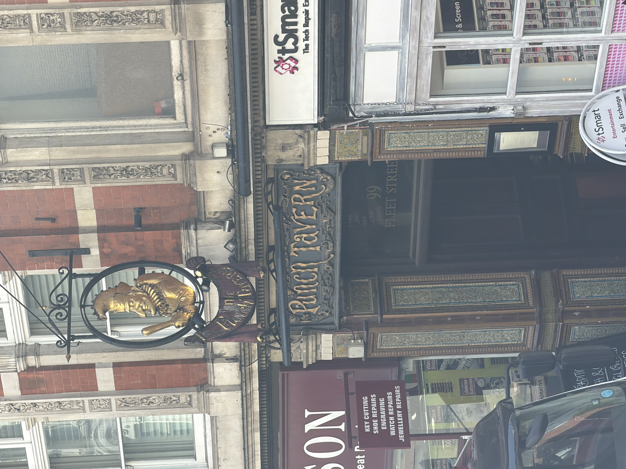 The image depicts the exterior of the Punch Tavern, located at 99 Fleet Street in London, United Kingdom. The building features intricate architectural details, including decorative moldings, brickwork, and window designs. A hanging sign with a golden sculpture of Punch and Judy characters is visible. Below the tavern sign, there is another sign for a repair shop, offering services like key cutting, shoe repairs, engraving, watch repairs, and jewelry repairs. The image also captures a glimpse of adjacent businesses, including a "tSmart" store. A vehicle is partially visible in the foreground, with its side mirror and parts of the car body reflected.