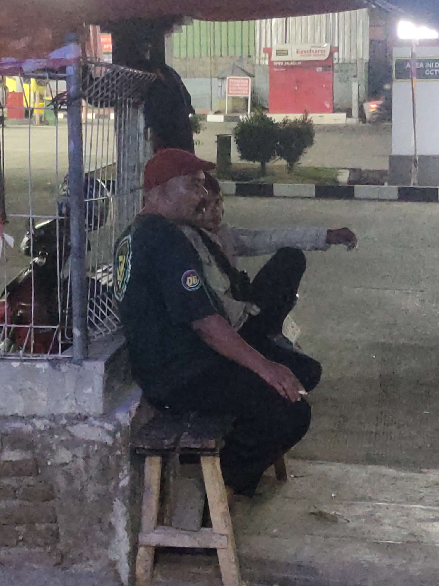 The image shows two individuals seated on a wooden stool in an outdoor urban setting, likely in Sumedang, Indonesia. The person in the foreground wears a red cap and a dark green t-shirt with a circular logo on the sleeve. They are holding a cigarette in their right hand and appear to be exhaling smoke. A second person, partially obscured and wearing a lighter-colored shirt and dark pants, sits next to them. A motorcycle is visible behind a metal fence to the left of the subjects. In the background, there are buildings with signage, including one that reads "Enduro." The ground is paved and appears to be a roadside or parking area. The overall lighting suggests it is either dusk or nighttime.
