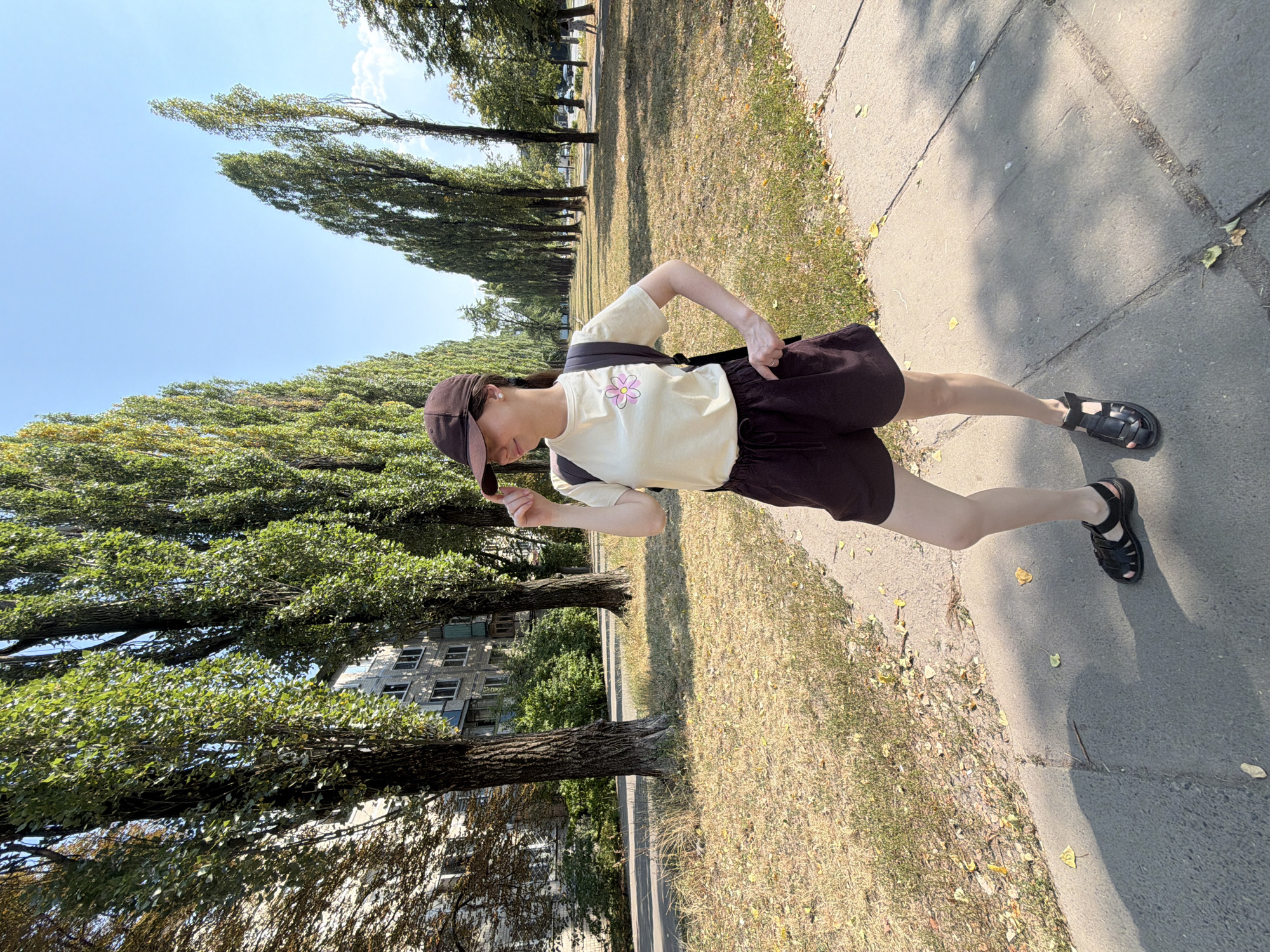 A young woman stands on a paved sidewalk in a park-like setting in Kyiv, Ukraine. She is wearing a light-colored t-shirt with a pink flower design, dark shorts, a dark baseball cap, and black sandals. She has a backpack slung over her left shoulder and is touching the brim of her cap with her right hand, looking to her right. The background features a row of tall, slender trees lining a path, with an apartment building visible on the left. The ground is covered in dry grass and fallen leaves, suggesting an autumn day. The sky is clear and blue.