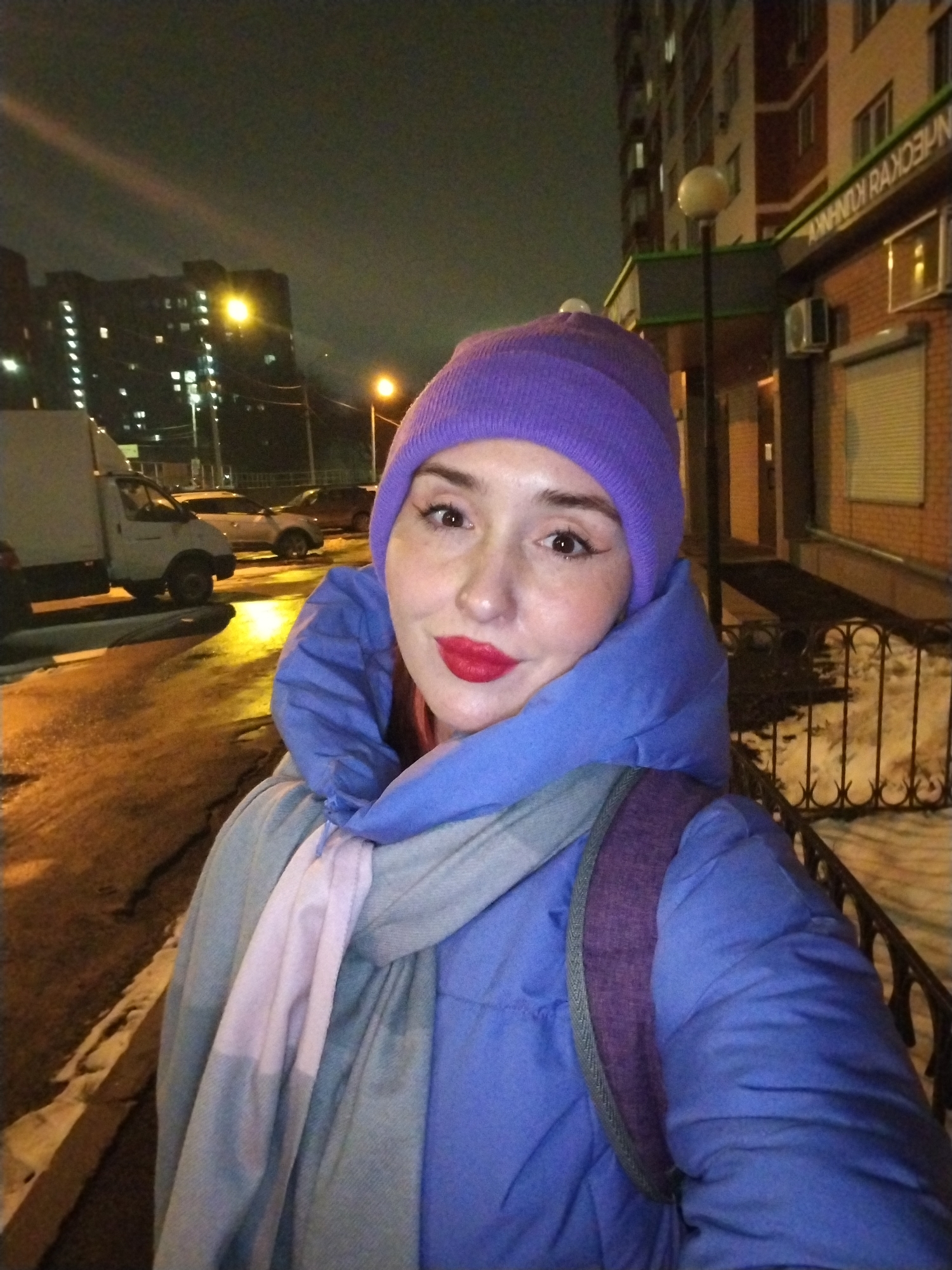 A young woman with fair skin and red lipstick is the focal point of the image. She is wearing a purple knit hat, a blue puffy jacket, and a grey scarf with a pale pink stripe. A purple backpack strap is visible over her shoulder.
She is standing outside at night, likely in a city. A light source illuminates the ground around her. Behind her to the left, there is a white van parked on a wet street, several other cars, and a large apartment building with lit windows. To her right, there is a brick building with signage in what appears to be the Russian language. A street light is positioned near the building, and a wrought-iron fence separates the building from a snow-covered area.
The location is identified as Izhevsk, Russia.
